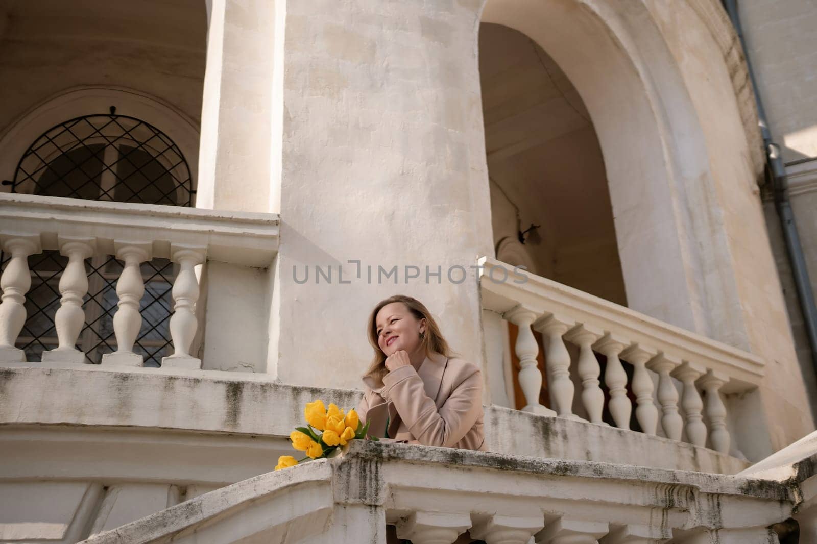 A woman wearing sunglasses and holding a bouquet of yellow flowers stands on a balcony. The scene is peaceful and serene, with the woman looking out over the city
