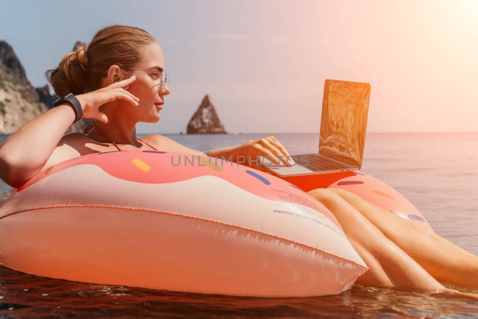 Woman freelancer works on laptop swimming in sea on pink inflatable ring. Pretty lady typing on computer while floating in the sea on inflatable donut at sunset. Freelance, remote work on vacation