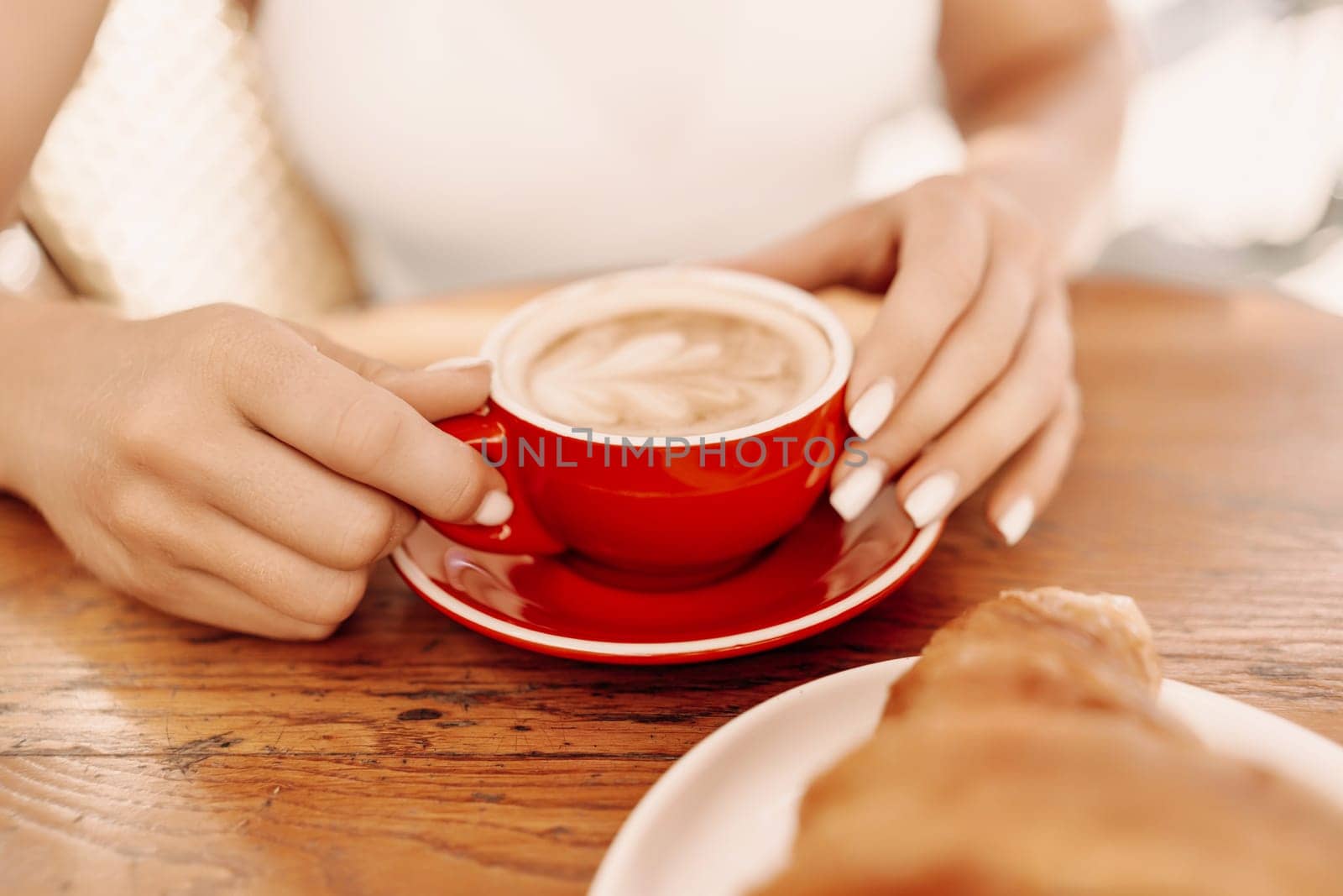 A woman is holding a red coffee cup with a white saucer. The woman is sitting at a table with a plate of croissants in front of her