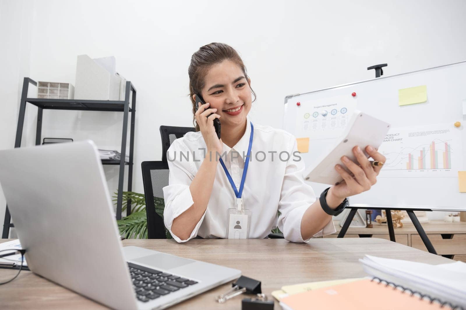 Asian female accountant is talking on the phone in the office with working documents regarding calculations on the office table. by wichayada