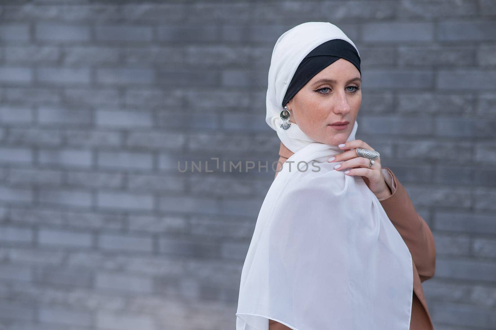 Portrait of a young blue-eyed woman in a hijab against a gray brick wall. A Muslim woman looks at the camera turning around