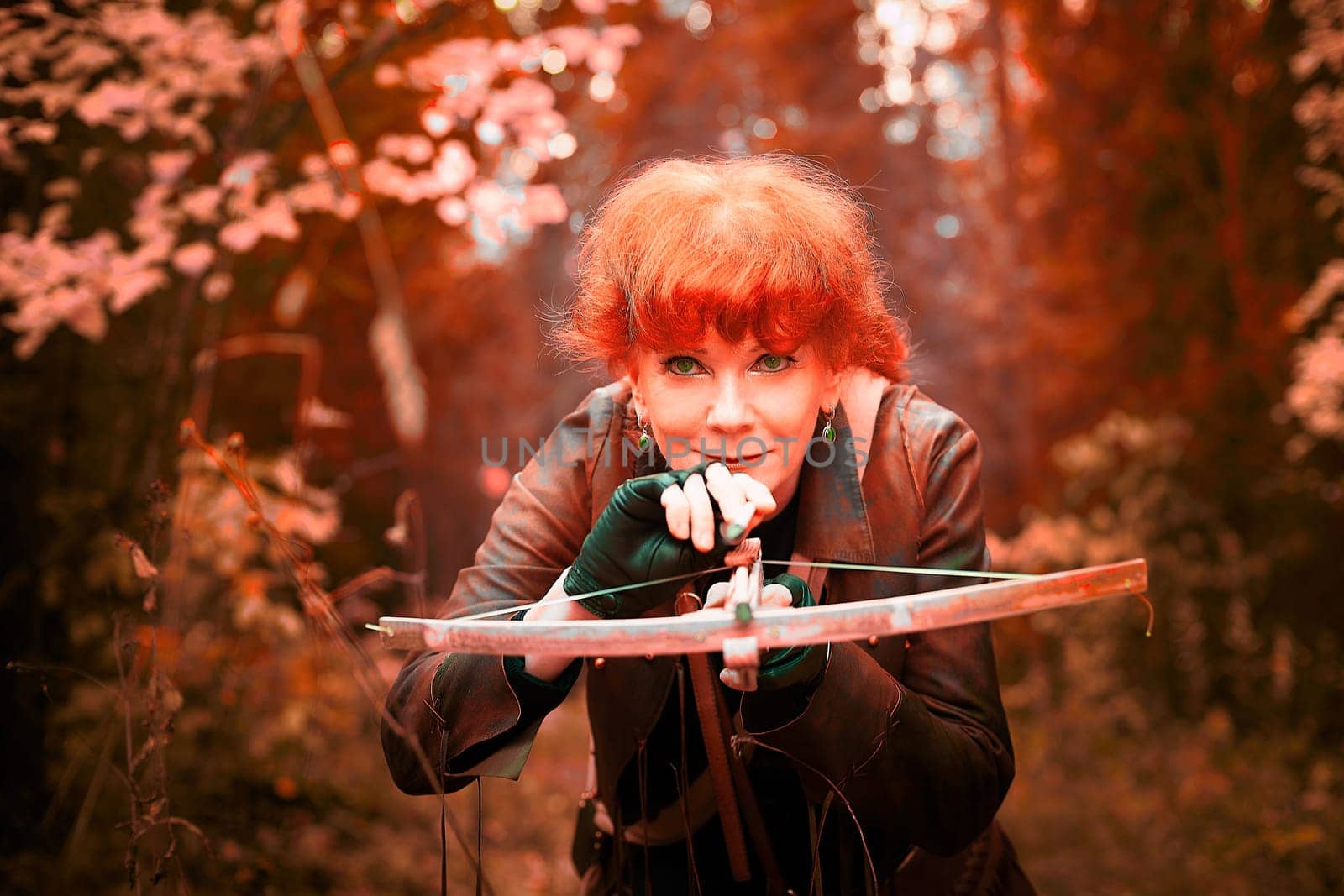 Mature model portraying a royal huntress with red curve hair is hunting with a crossbow in the in vibrant autumn forest in thematic photo shoot
