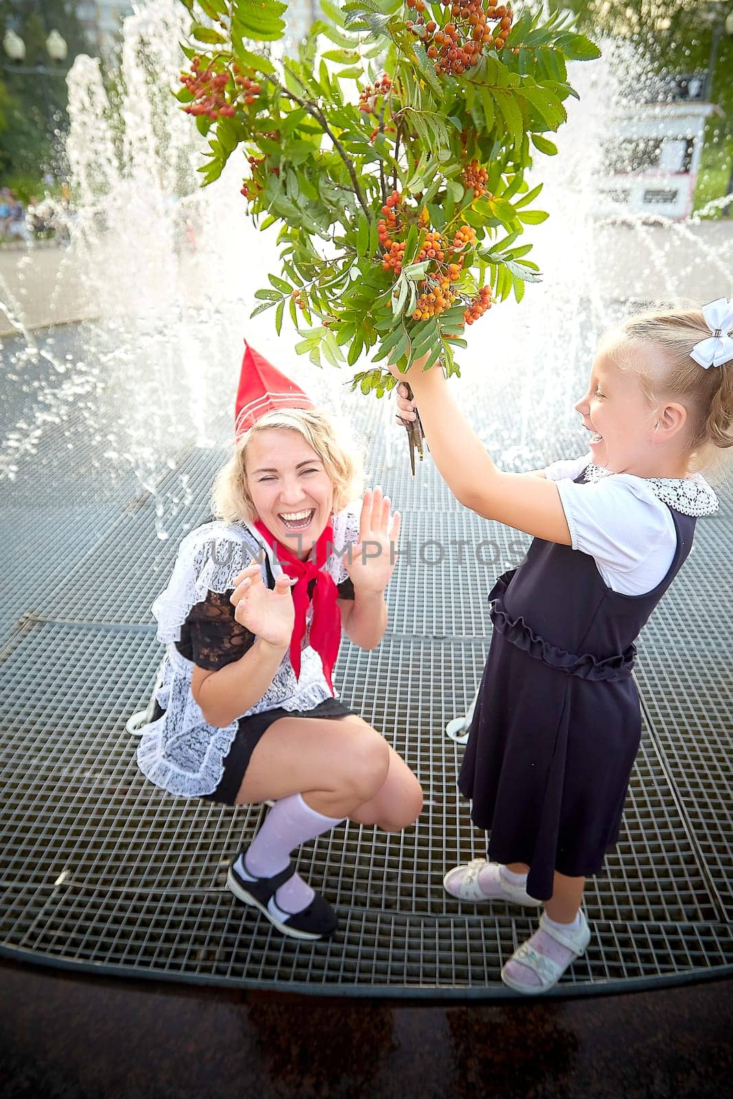 Young and adult schoolgirl on September with flowers having fun near water of fontain. Generations of schoolchildren, pioneer of USSR and October girl in modern uniform of Russia. Mom and daughter