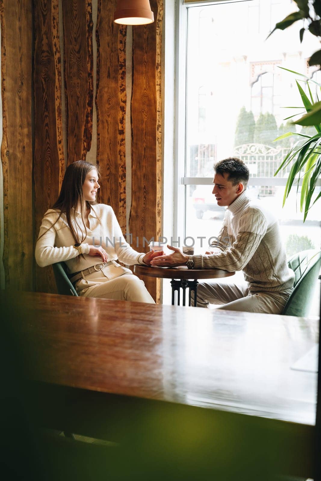 Two professionals are engaged in a conversation while sitting opposite each other at a wooden table in a cafe with natural light streaming in from a large window. They appear relaxed and focused, with cups of coffee in hand, surrounded by a warm, rustic interior.
