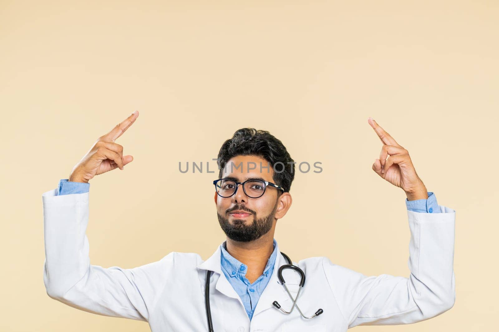 Indian young doctor cardiologist man show thumbs up and pointing overhead empty place advertising area for commercial discount, copy space. Arabian apothecary pharmacy guy isolated on beige background