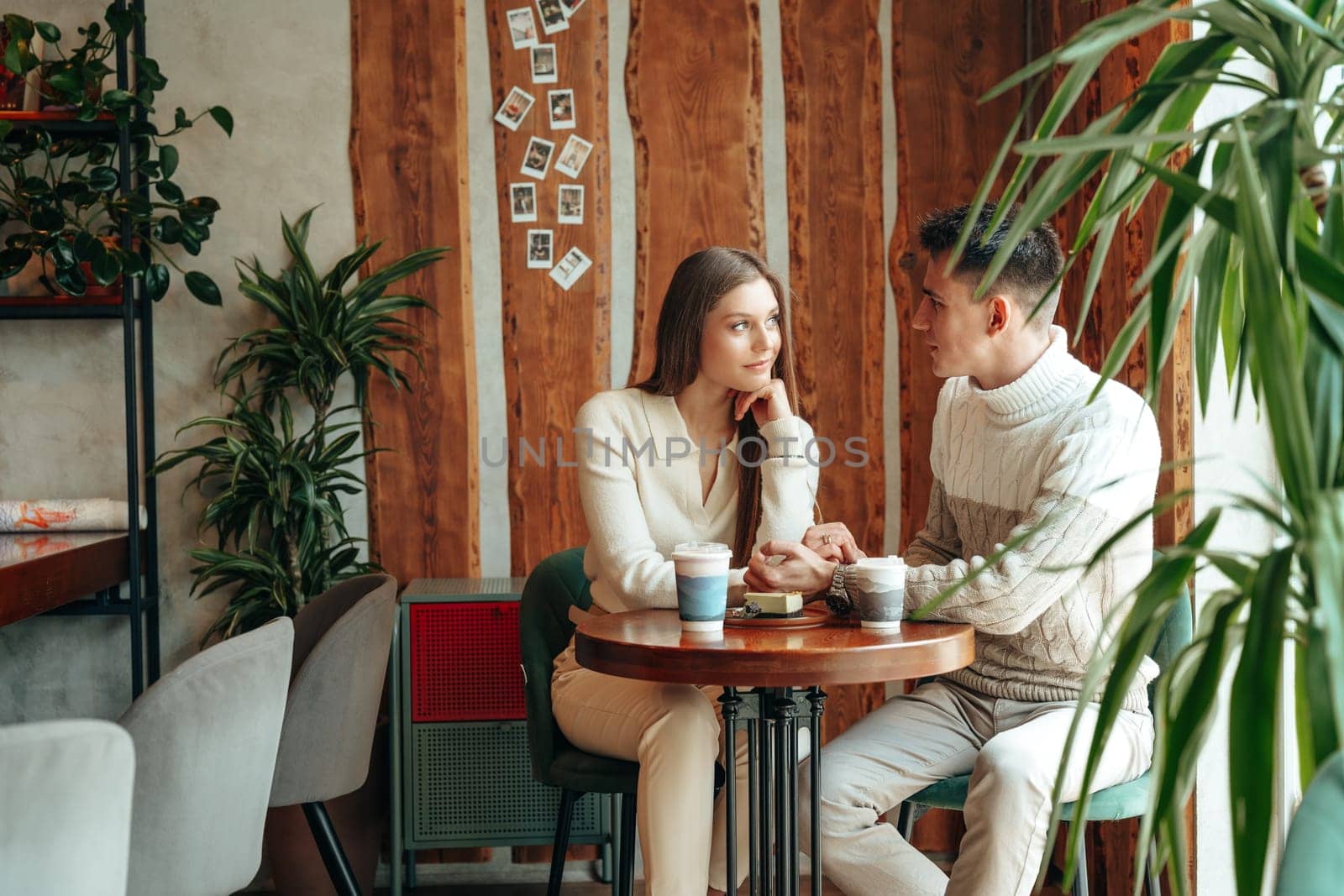 A young man and woman are seated across from each other at a small wooden table in a charming cafe, engrossed in a serene conversation. The woman is resting her chin on her hand, listening intently, while the man gestures as he speaks. The warm, casual atmosphere is enhanced by the presence of indoor plants and decorative items on the wall, suggesting a leisurely daytime outing.