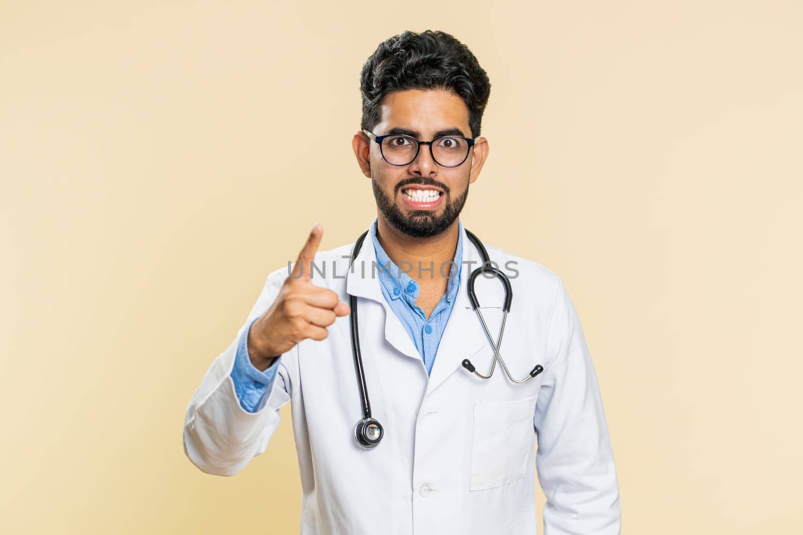 Indian young doctor cardiologist man shakes finger and saying no, be careful, advice to avoid danger mistake, disapproval sign, disagree, dislike. Arabian apothecary pharmacy guy on beige background