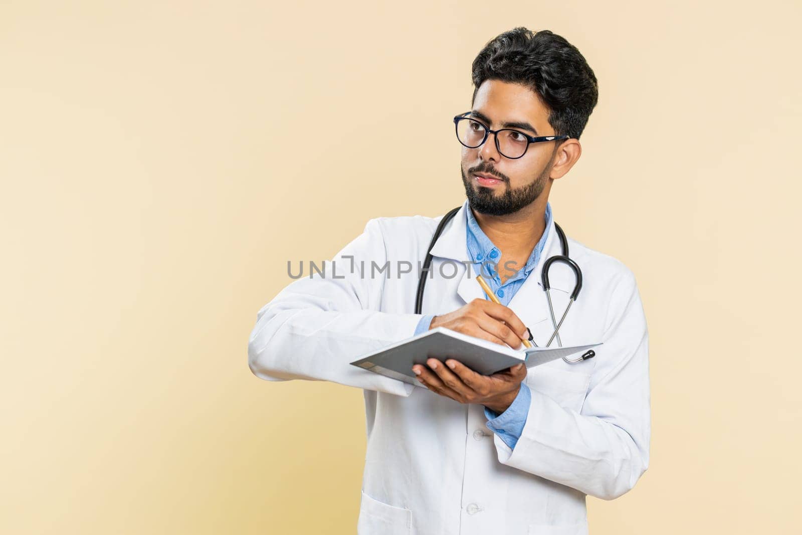 Thoughtful Indian young doctor cardiologist man making notes, writing down thoughts with pen into notepad notebook diary, to do list, good idea. Arabian apothecary pharmacy guy on beige background