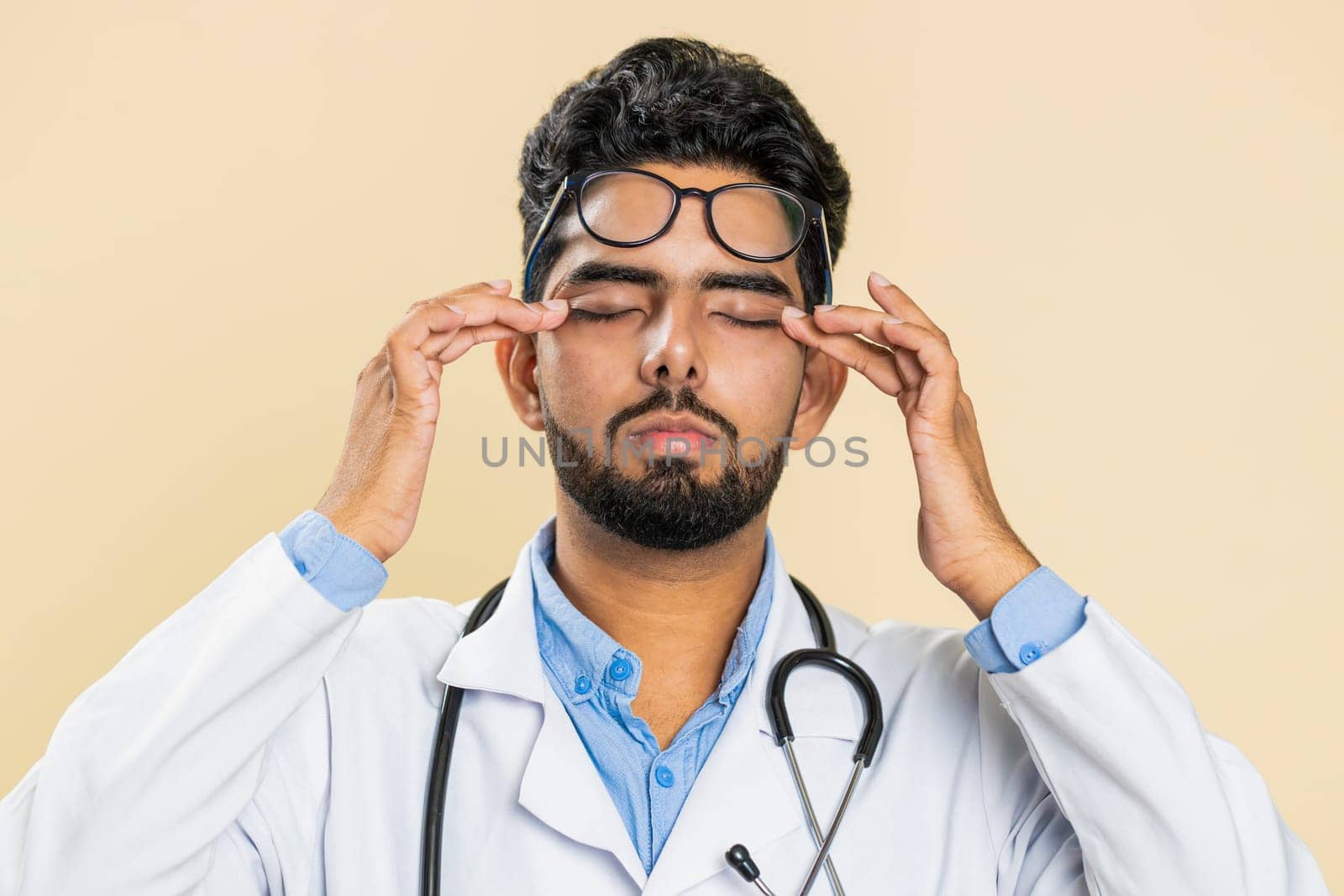 Exhausted tired Indian young doctor cardiologist man takes off glasses, feels eyes pain, being overwork burnout from long hours working. Sleepy exhausted guy rubbing eyes isolated on beige background