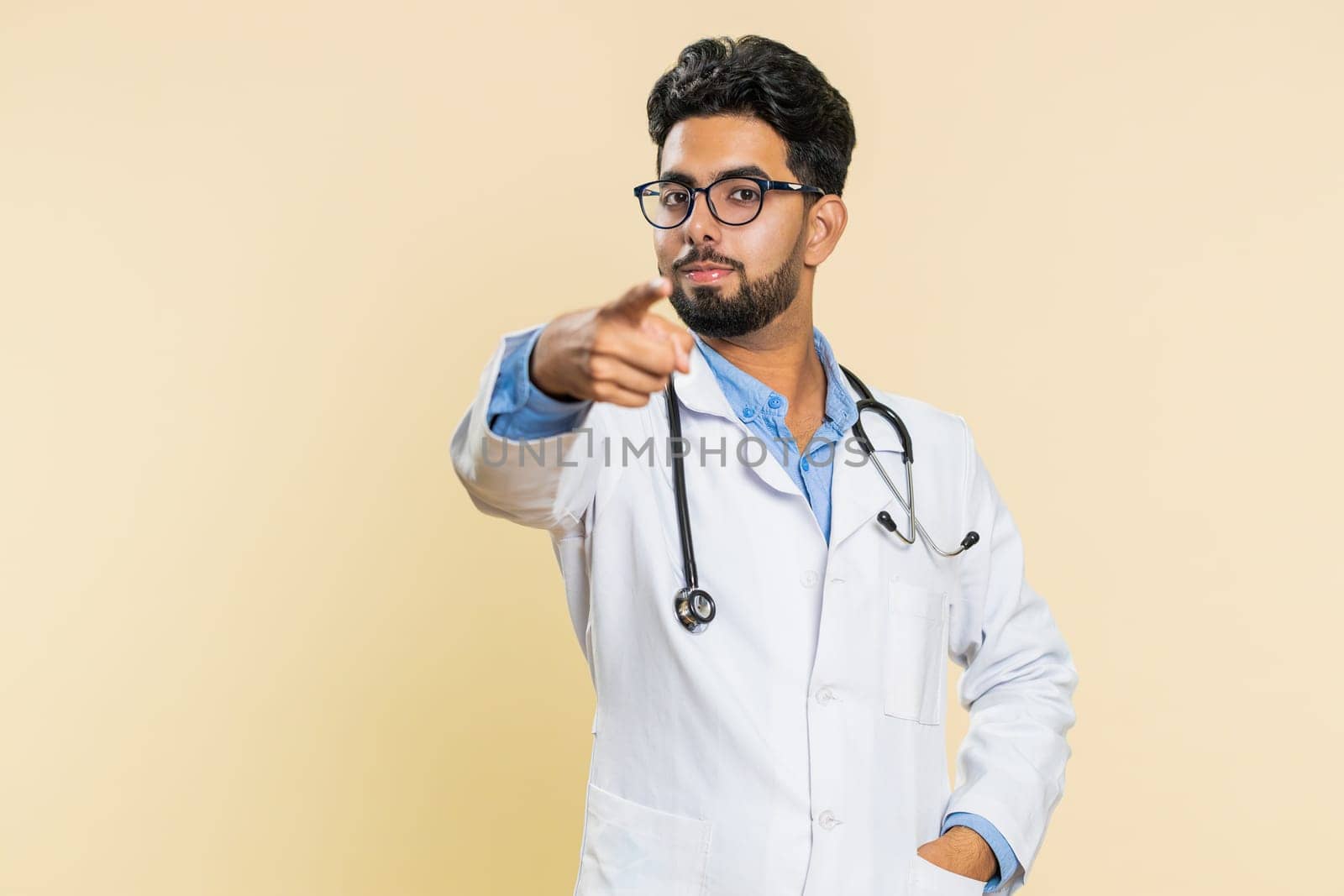 Hey you. Happy strict Indian young doctor cardiologist man pointing forward to camera, choosing, indicating to you, promotion. Arabian apothecary pharmacy smiling guy isolated on beige background