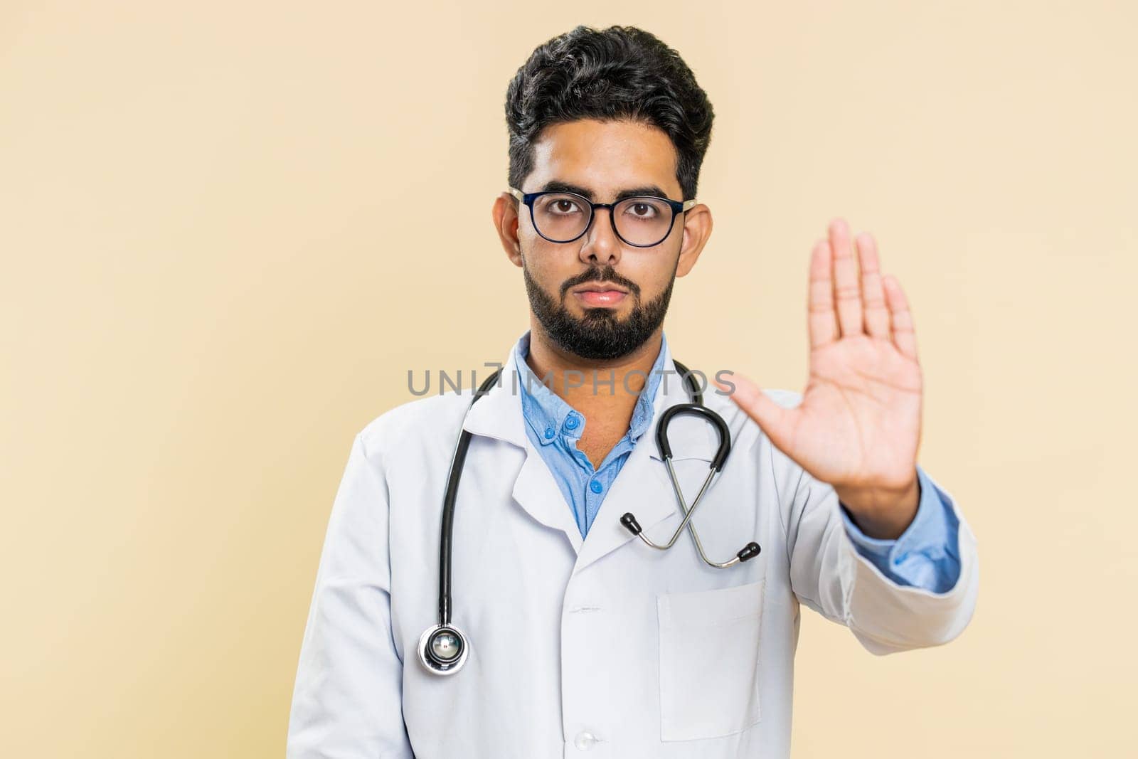 Stop, enough. Serious Indian young doctor cardiologist man say no, warning of finish, prohibited access gesture, declining communication. Arabian apothecary pharmacy guy isolated on beige background