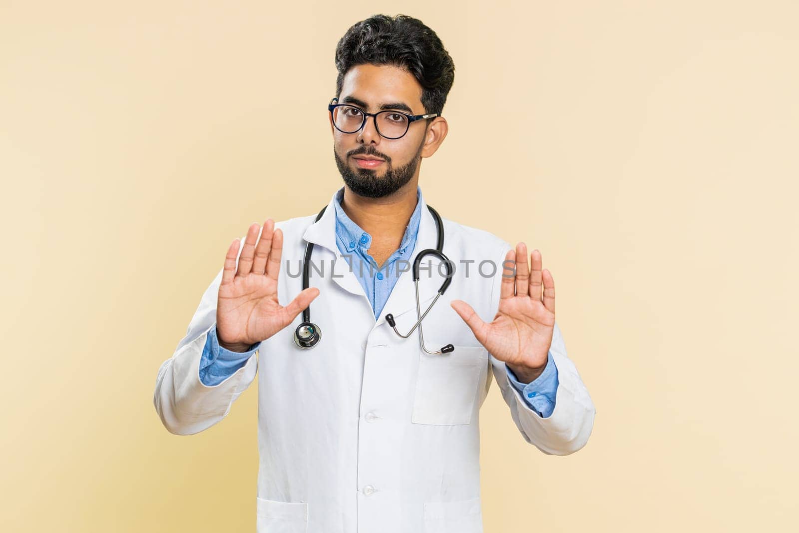 Hey you, be careful, take care. Strict Indian young doctor cardiologist man warning with admonishing hands gesture, giving advice to avoid danger. Arabian apothecary pharmacy guy on beige background