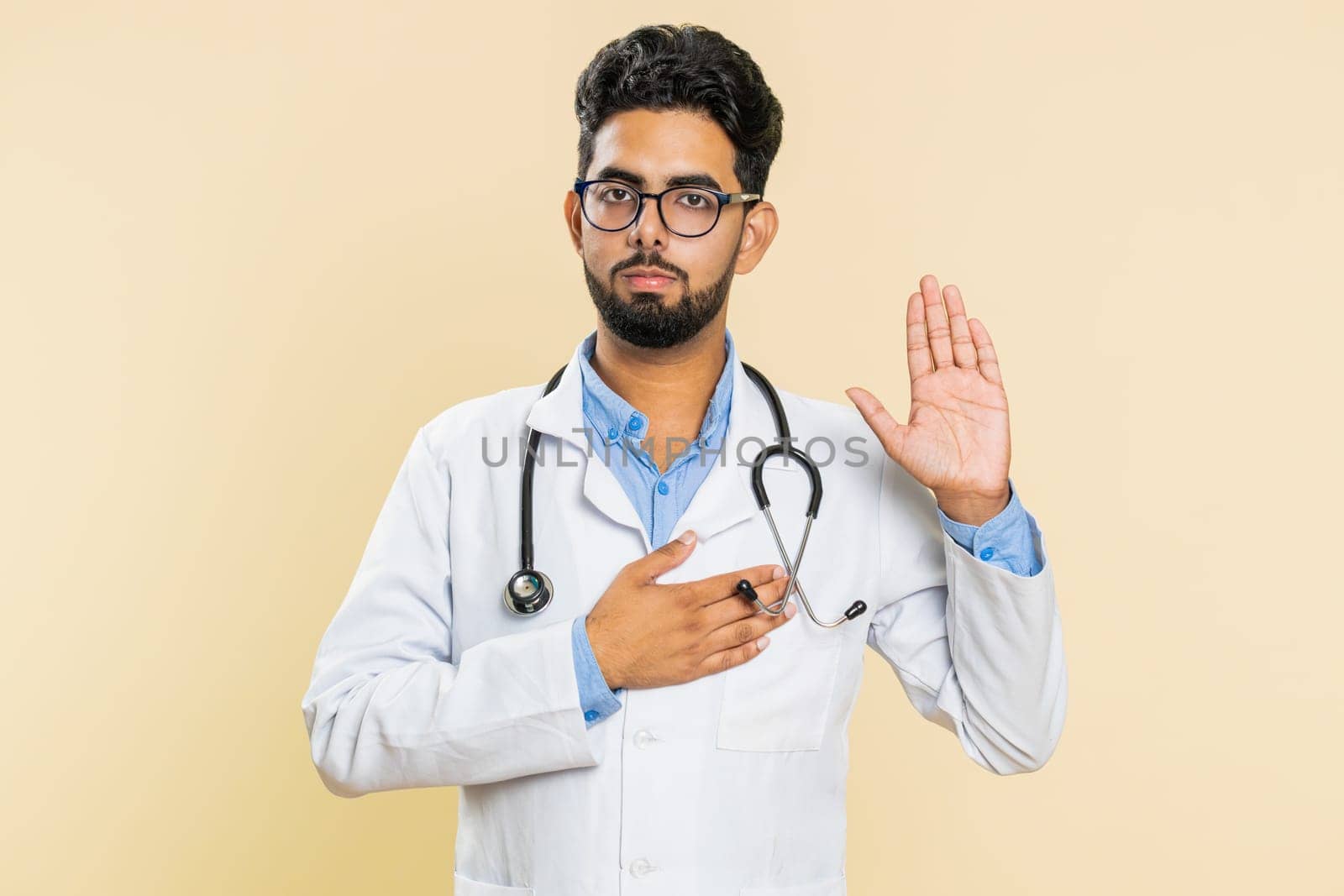 I swear to be honest. Sincere responsible Indian young doctor cardiologist man raising hand to take oath, promising to tell truth, keeping hand on chest. Apothecary pharmacy guy on beige background