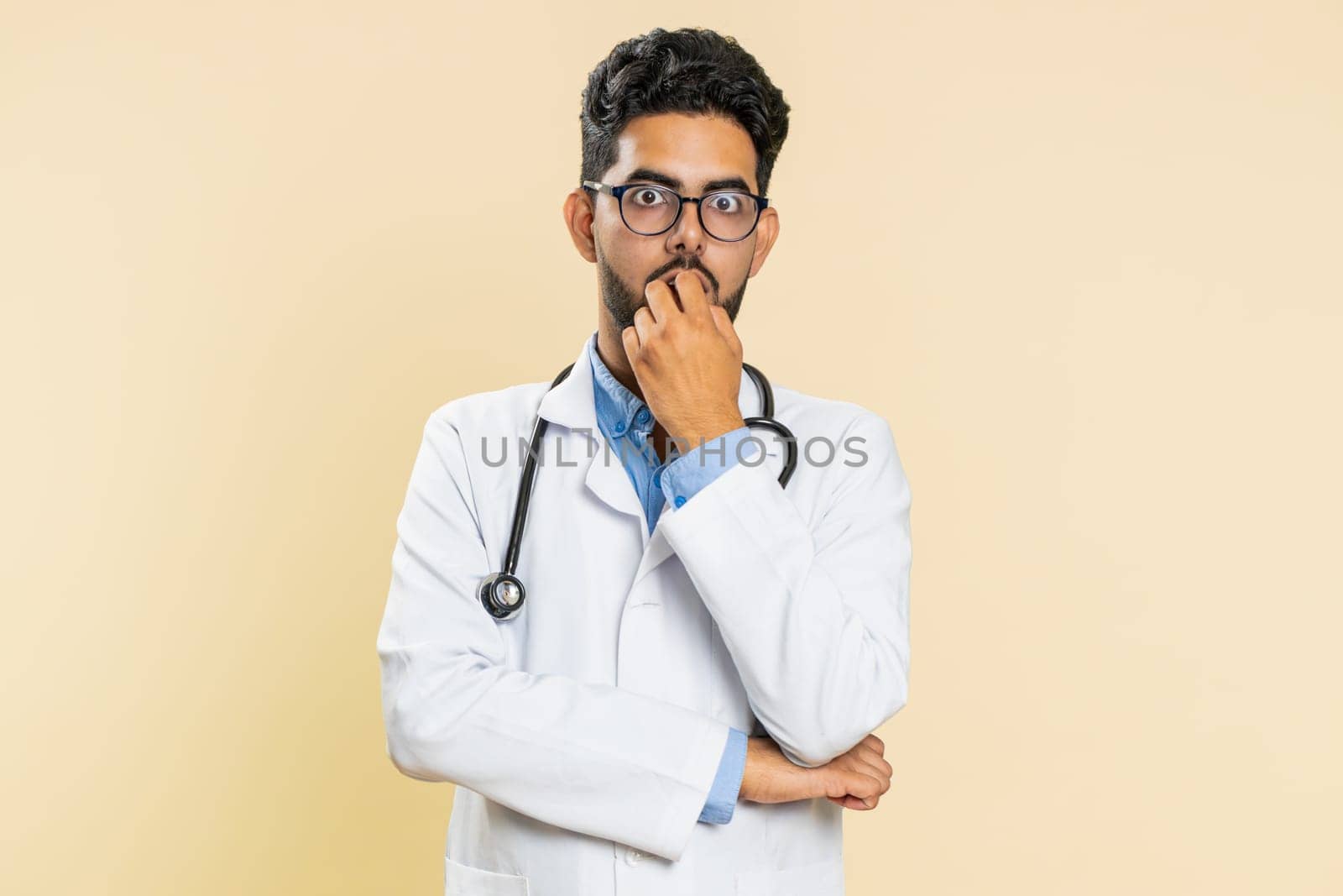 Upset scared frightened Indian doctor cardiologist man biting nails, feeling worried nervous about serious troubles, stress, anxiety disorder, panic attack, guilty. Arabian guy on beige background