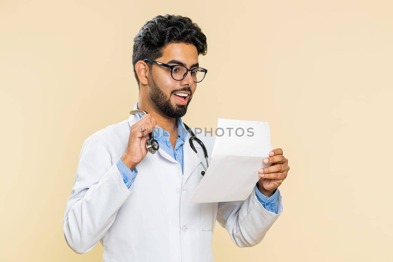 Indian young doctor cardiologist man open envelope take out letter reads it feel happy. Career growth advance promotion, long-awaited invitation great news. Arabian apothecary guy on beige background