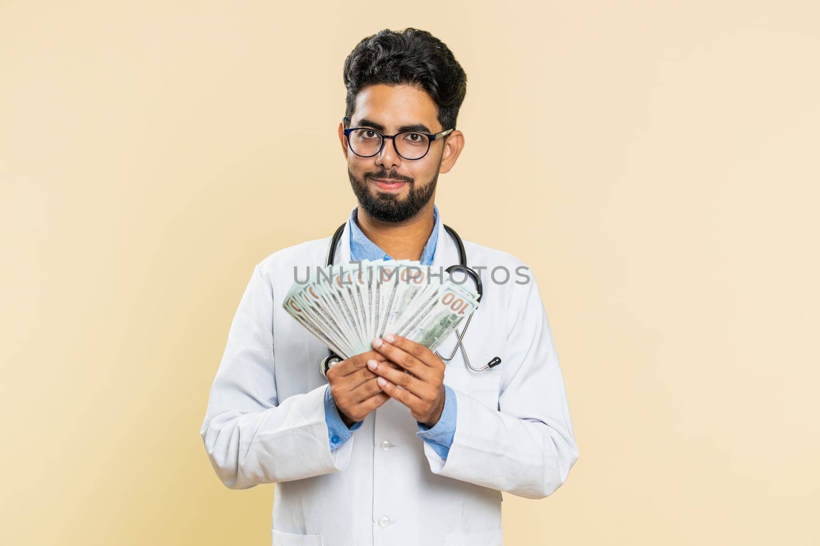 Rich happy Indian doctor cardiologist man waving money dollar cash banknotes bills like a fan, success business career, big income wealth. Arabian apothecary pharmacy guy isolated on beige background