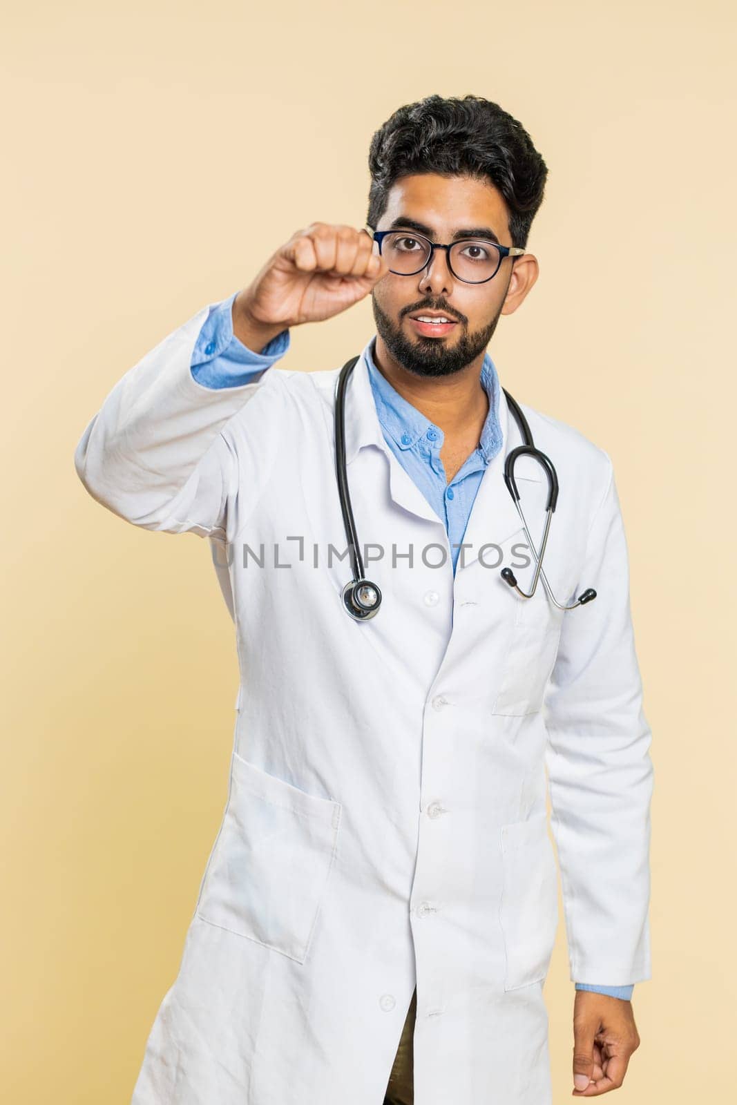 Knock-knock who is there. Excited Indian doctor cardiologist man knocking door gesture asking who is at home, being clueless uncertain. Hello, welcome. Arabian apothecary on beige background. Vertical