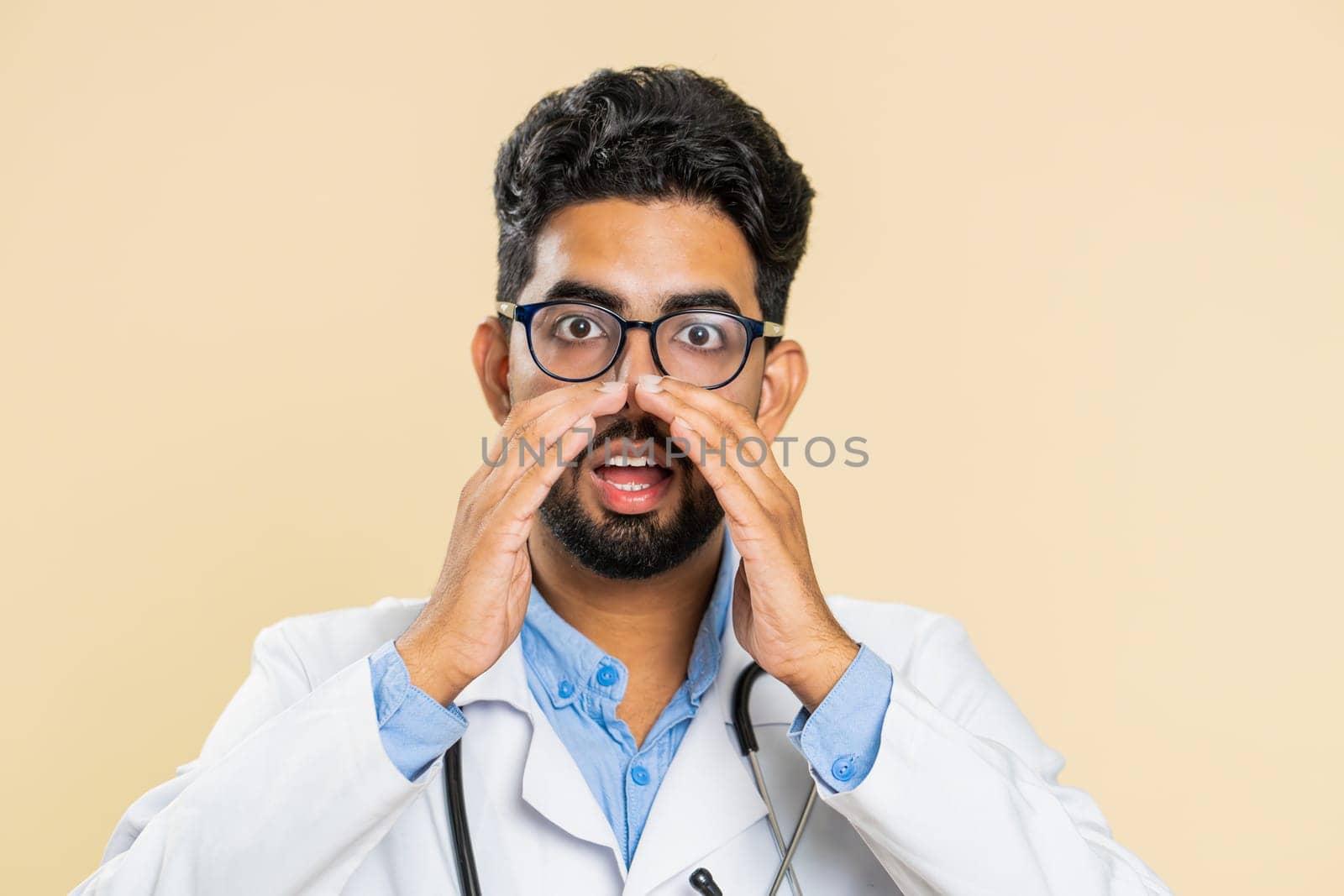 Keep my secrets, silence. Indian doctor cardiologist man whisper news rumors holding hands near mouth, share gossip, quiet. Arabian guy telling interesting confidential information on beige background