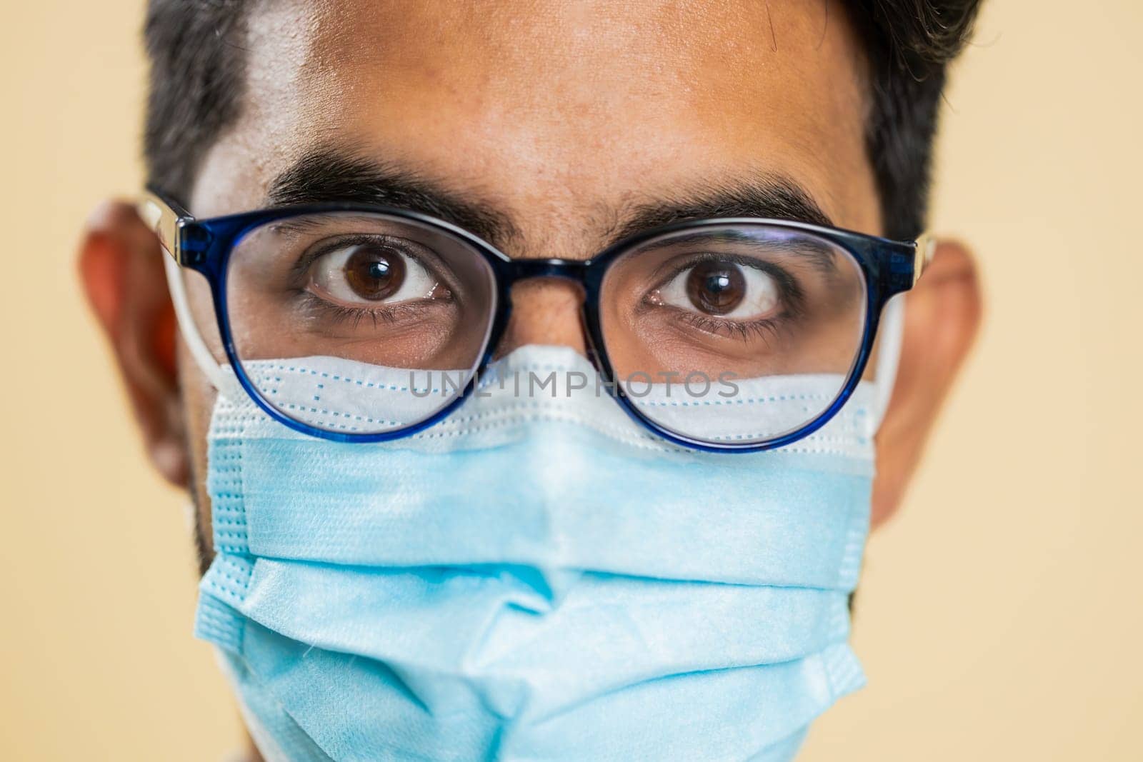 Extreme close up face portrait of Indian young doctor cardiologist man lab worker in glasses looking at camera. Arabian apothecary pharmacy guy in medical protective mask isolated on beige background