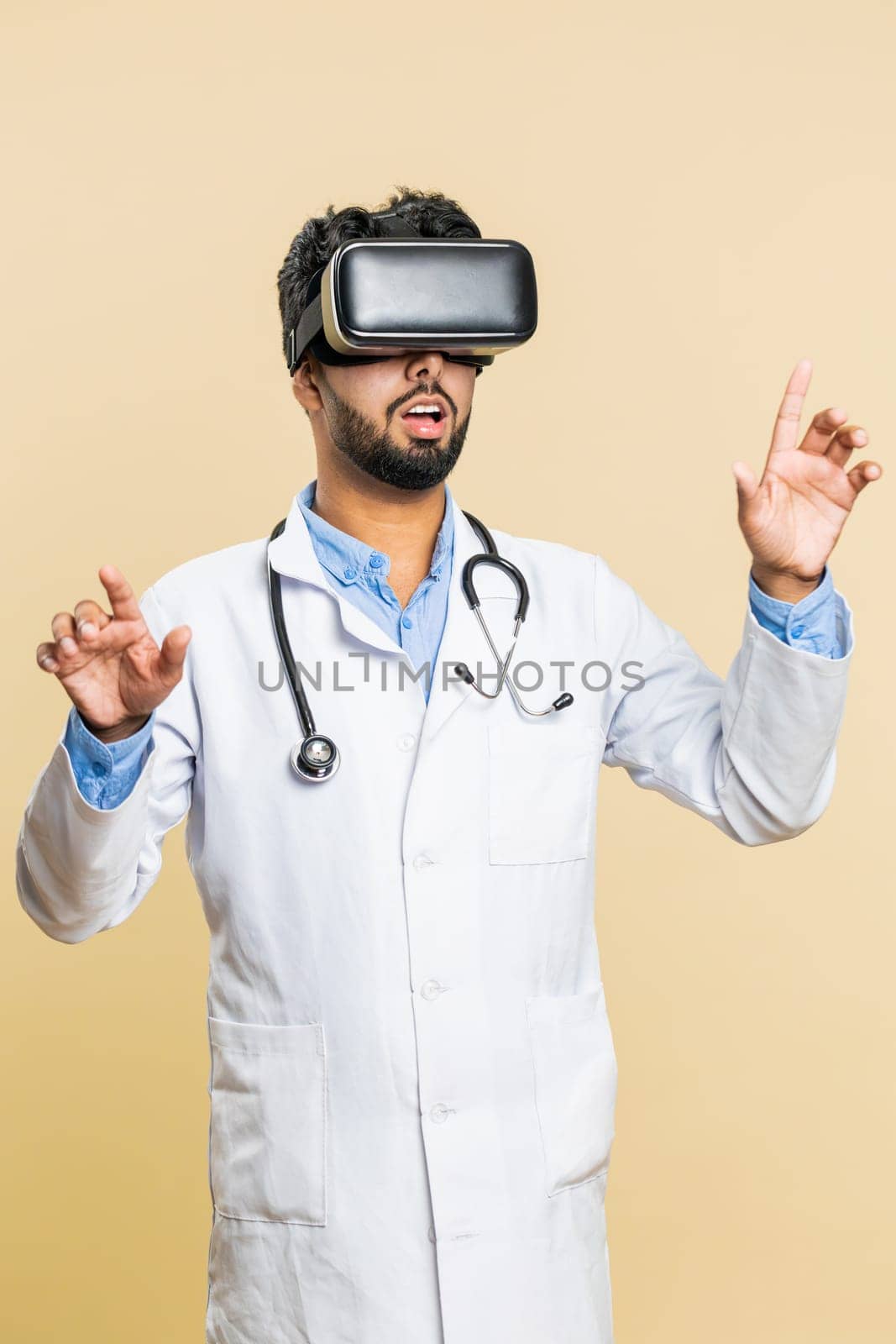 Excited Indian doctor cardiologist man using headset helmet app, watching virtual reality 3D 360 video. Arabian apothecary guy in VR goggles on beige background. Future medicine technology. Vertical