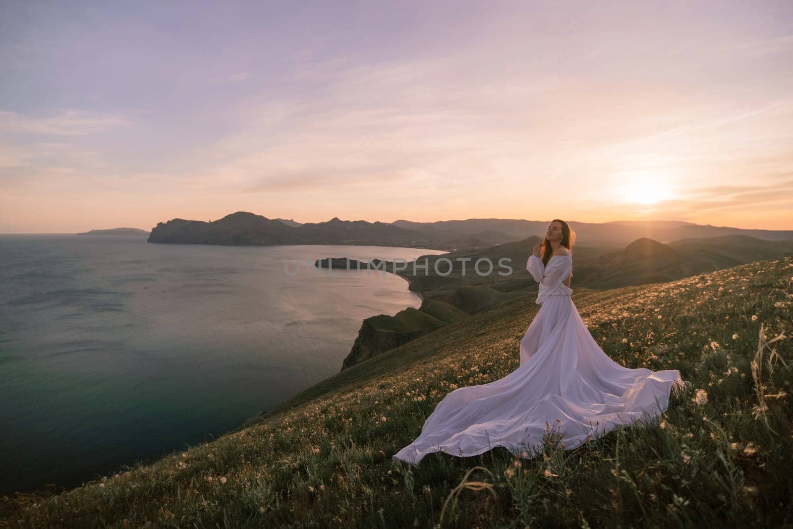 A woman in a white dress stands on a hill overlooking a body of water. The scene is serene and peaceful, with the sun setting in the background. The woman is lost in thought