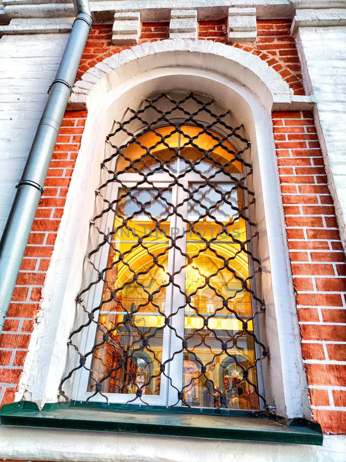 Warm light spills from the stained glass of a grilled window. Ornate Grille on Window of Brick Building at Dusk by keleny