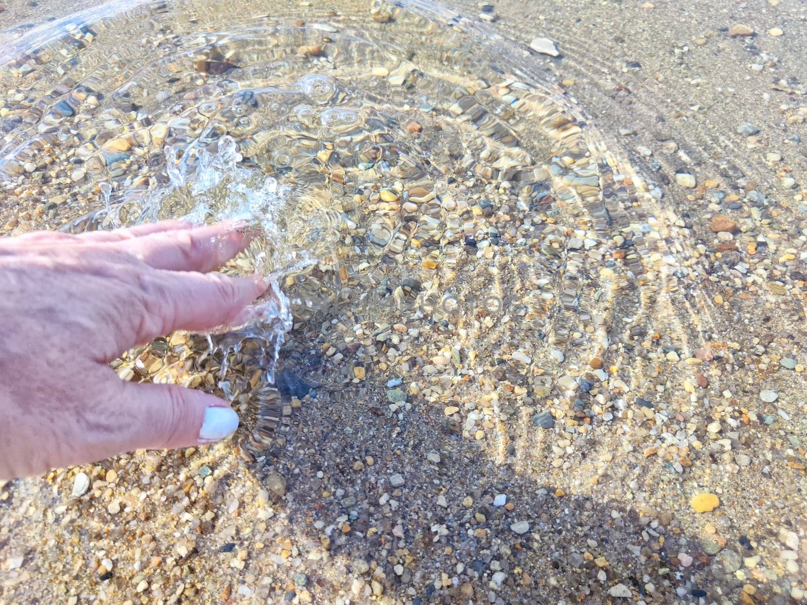 Pebbles in the water. Background, texture. Hand Disturbing Clear Water Over Colorful Pebbles. A hand touching clear water above pebbles