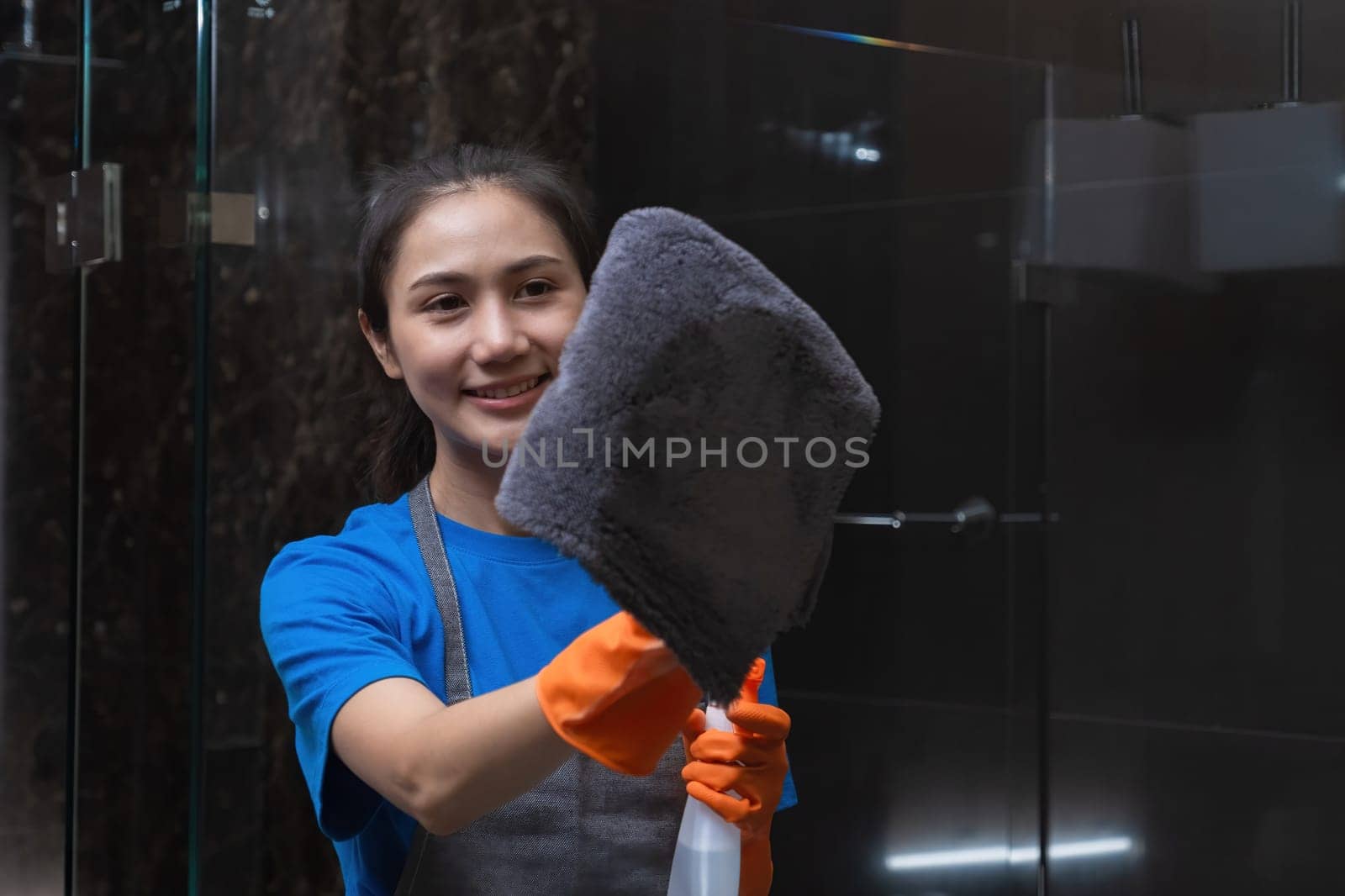 Beautiful housewife wearing orange cleaning gloves uses spray and cloth to clean the clear glass in the room. by wichayada