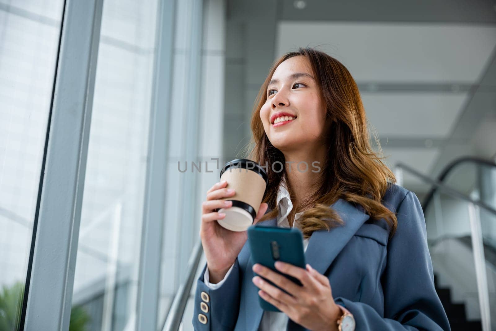Young Asian businesswoman wearing blue shirt holding coffee cup and her smartphone. She smiling and looking out of the window and elevator blur background in the office.