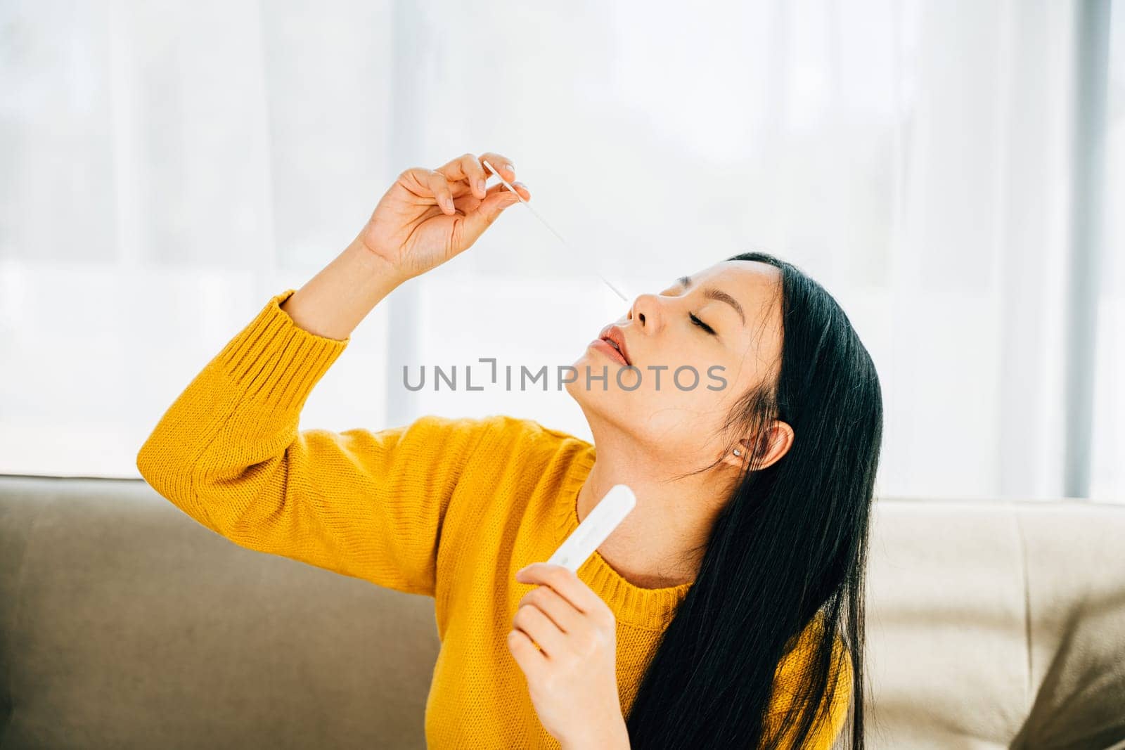 Highlighting COVID-19 self-test, Asian woman uses a rapid antigen test kit at home inserting a swab into her nose. Depicting virus prevention and pandemic protection measures.