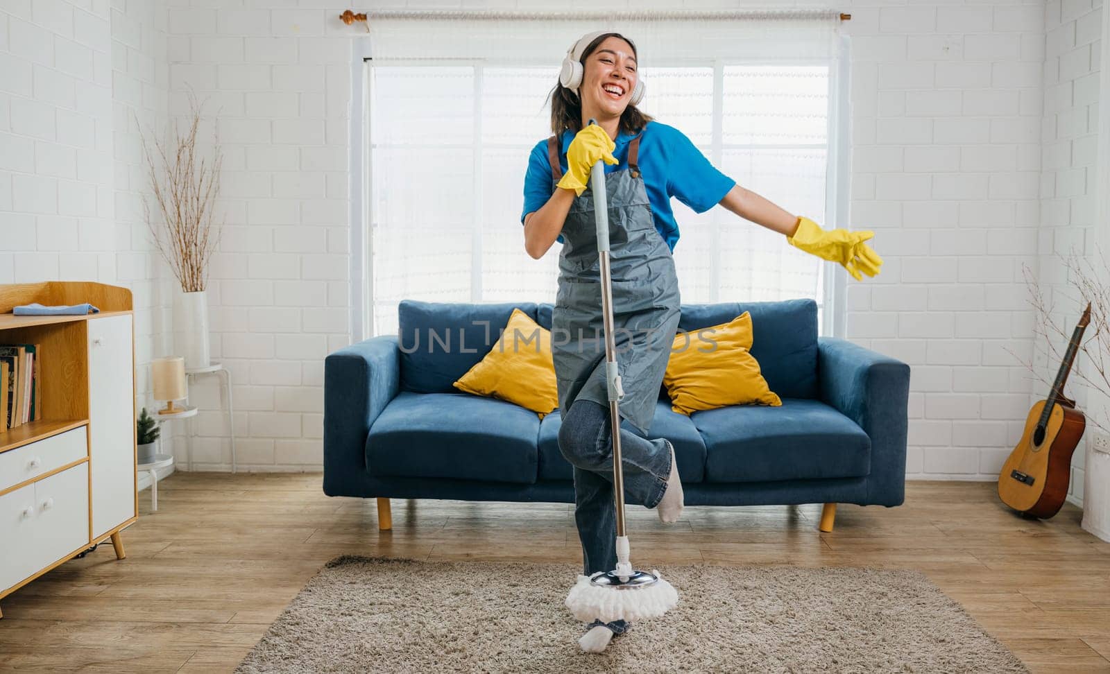 Happy young housewife enjoys singing her favorite song with mop as microphone during cleanup making housework fun. Dancing and cleaning in living room cheerful maid singing and enjoying her chores.
