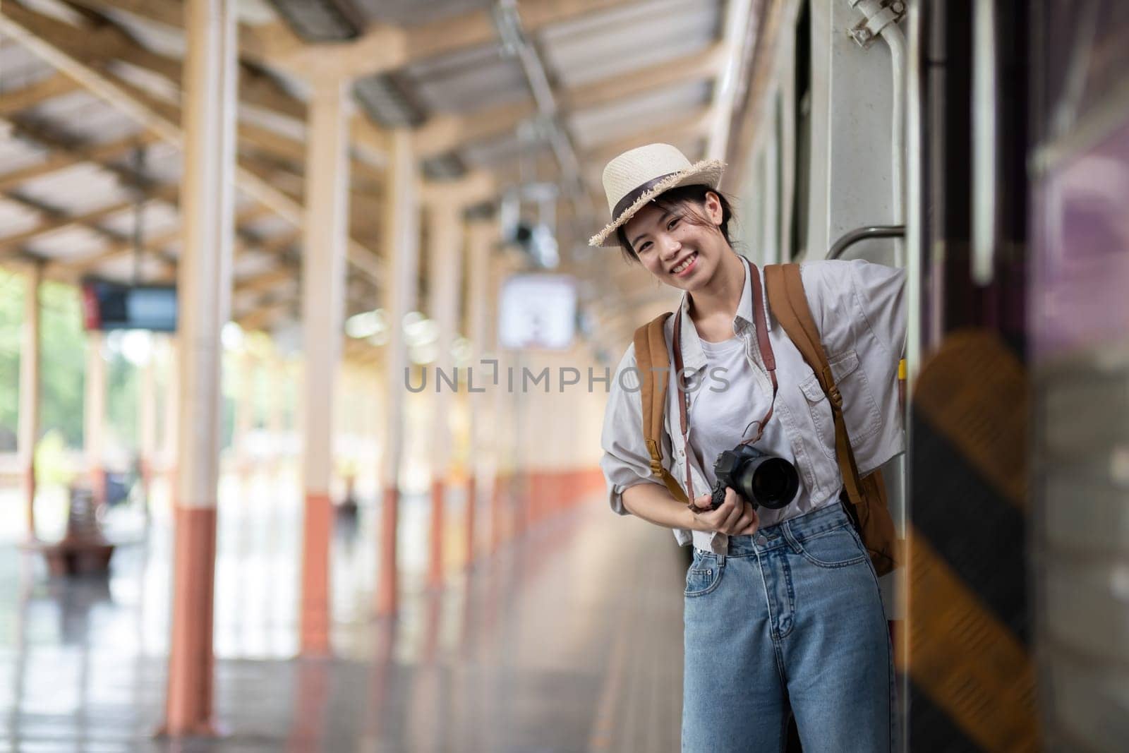 Happy young Asian female tourist carrying a backpack, holding a camera, preparing to wait for the train at the train station waiting for her vacation trip..