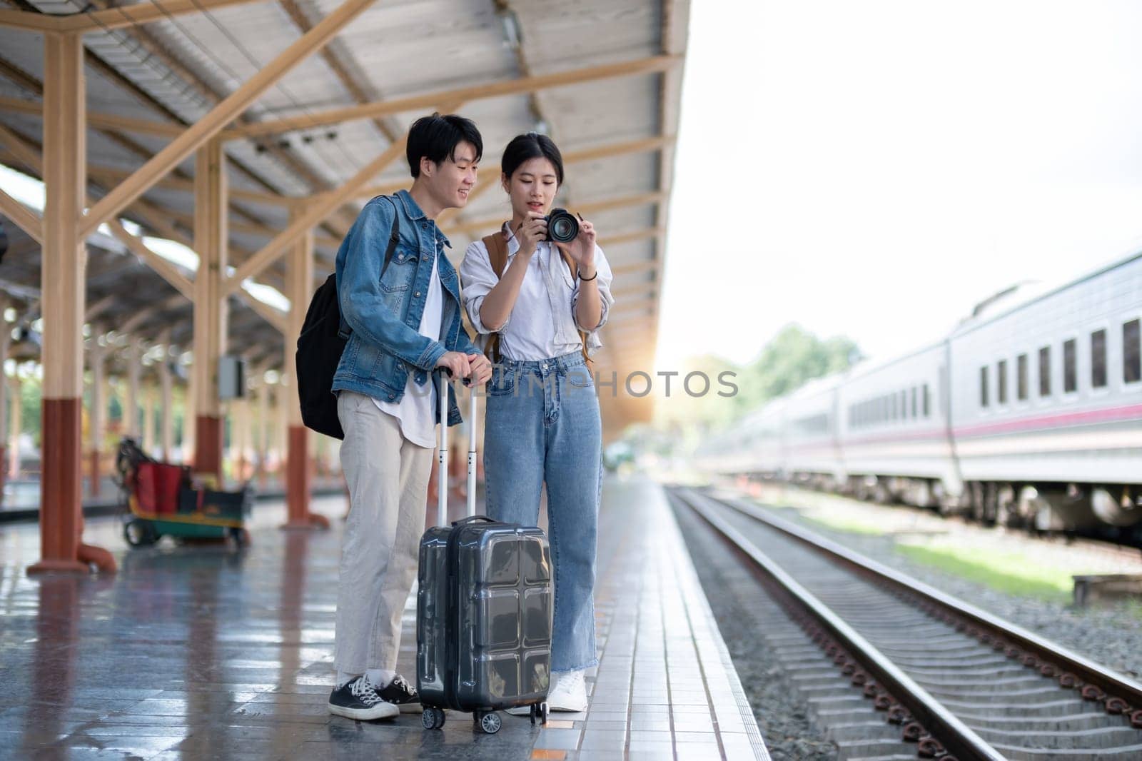 Happy young Asian couple carrying backpacks and cameras preparing to wait for the train at the train station waiting for their vacation trip together..