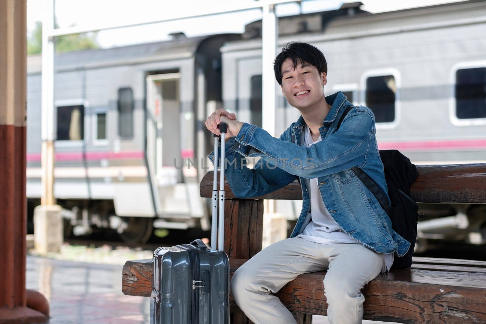 A young man holding a suitcase waits for a train at the train station for traveling. by wichayada