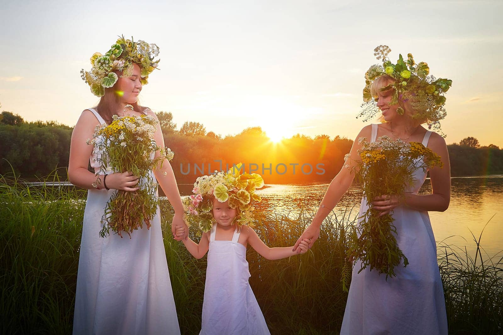 Ivan Kupala Celebration. Three Girls With Floral Wreaths by the River at Sunset. Family clad in white dresses celebrate Ivan Kupala by a river at dusk. by keleny