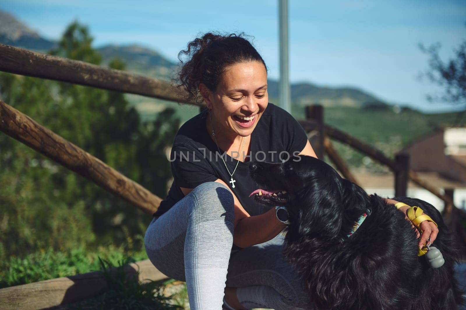 Happy smiling multi ethnic young woman hugging her pet dog, standing together on the forest path. Pretty woman walking her dog on the nature