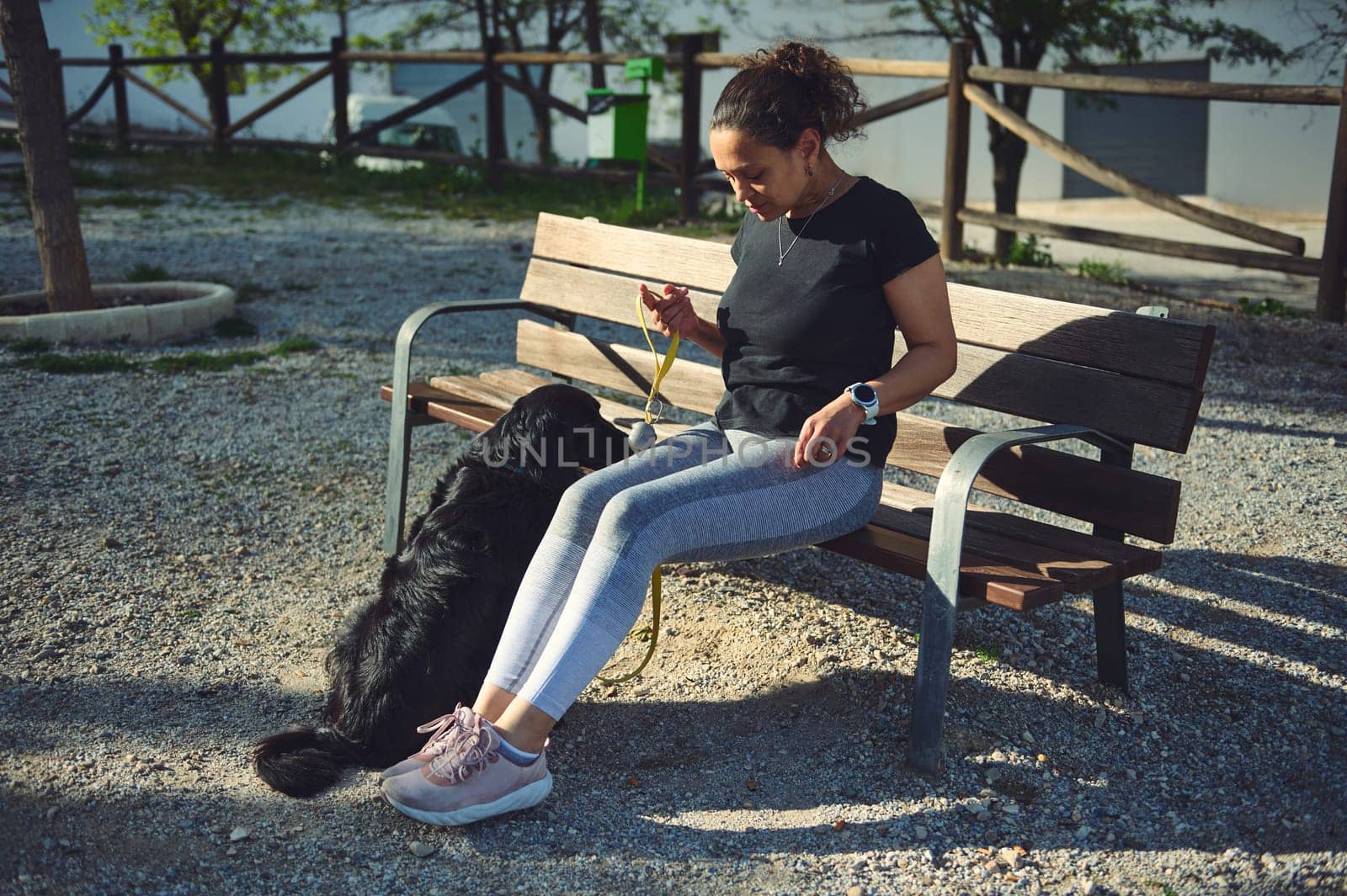 Young adult athletic woman relaxing sitting on the bench, walking her dog while jogging in the morning in the nature. Pets concept. People and domestic animals. Healthy active lifestyle concept