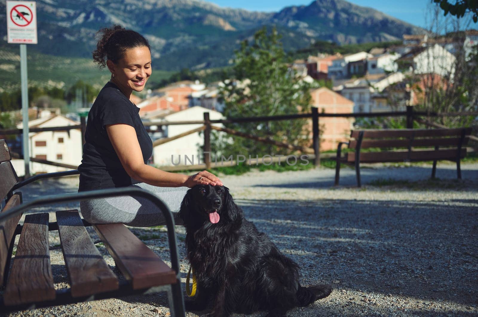 Happy young woman stroking her dog being walked on leach on the nature, sitting on the bench against mountains background. Pets concept. People and domestic animals. Healthy active lifestyle concept