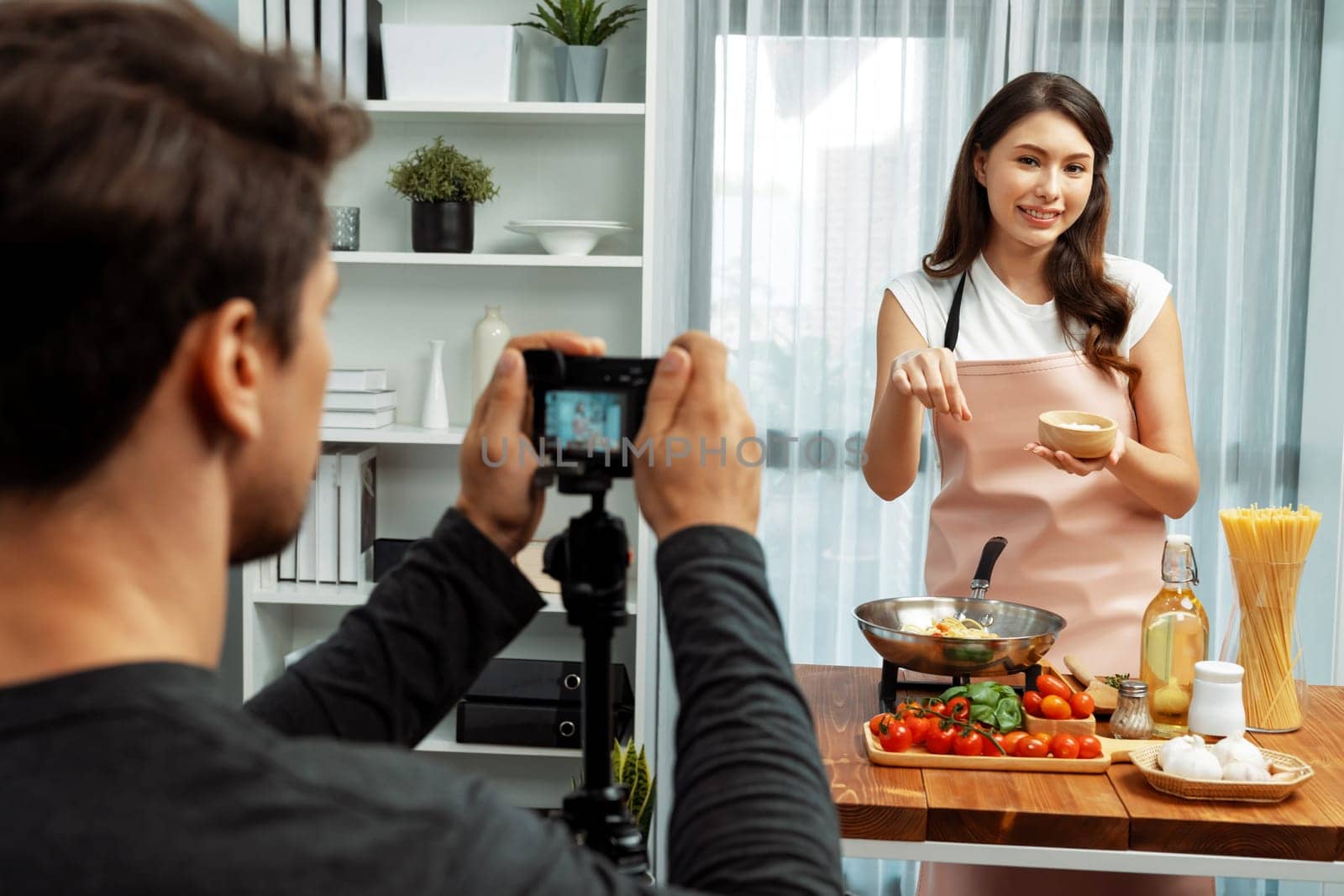 Cameraman recording to woman in chef influencer host cooking spaghetti with meat topped tomato sauce surrounded ingredients recipe, presenting special dish healthy food at modern studio. Postulate.