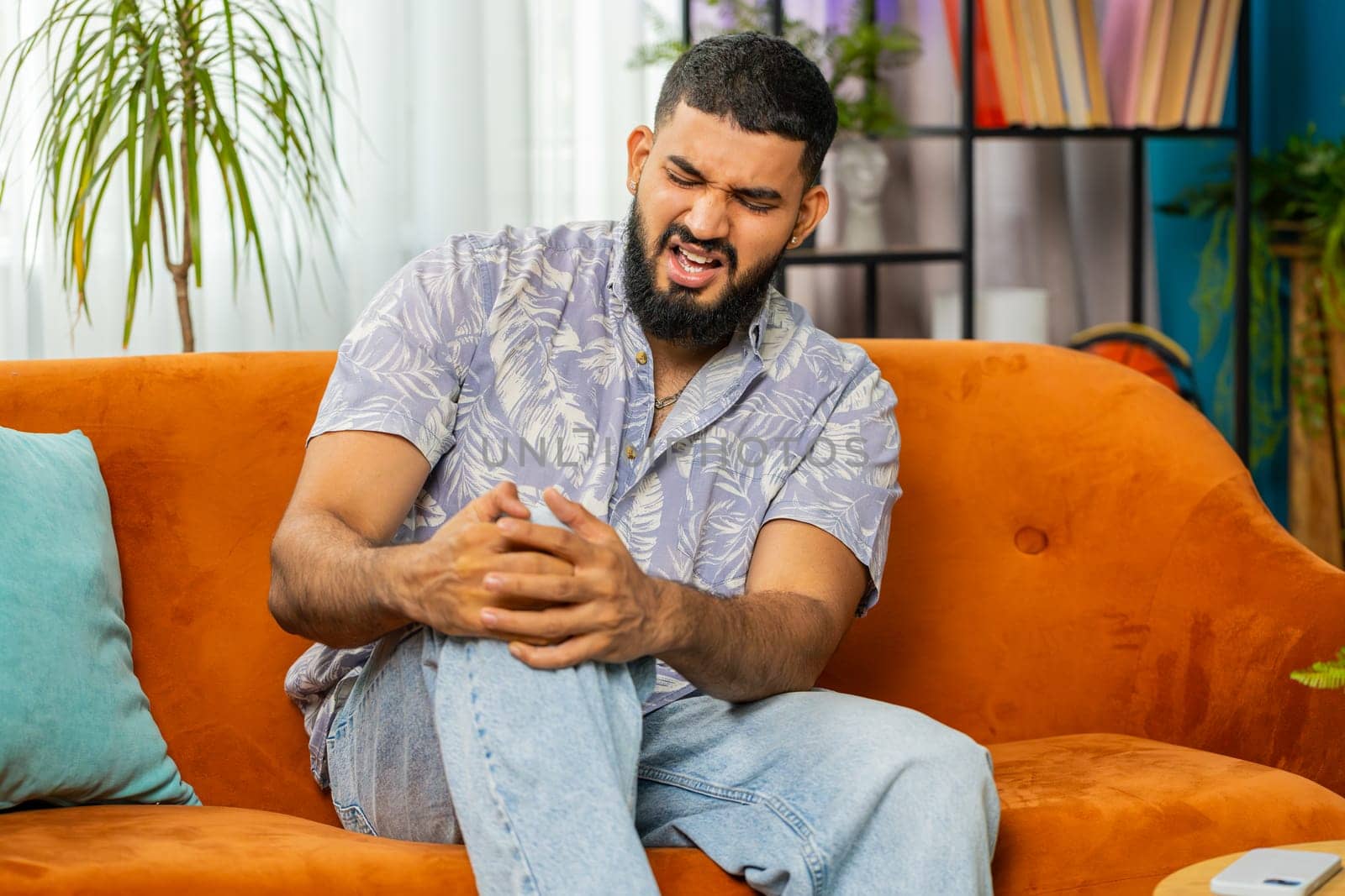 Indian man suffer from painful discomfort severe knee joint ache, leg muscles, could not walk. Sick Arabian guy massaging knees illness cramps rheumatism at home room on sofa. Health care problem