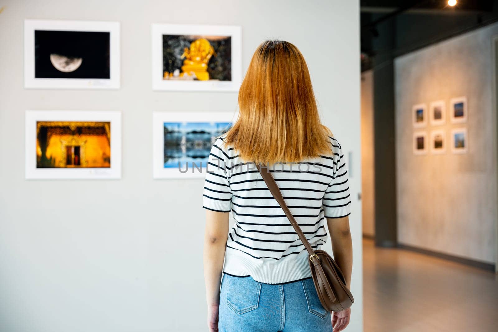Asian woman standing she looking art gallery in front of colorful framed paintings pictures on white wall, young female watch at photo frame to leaning against at show exhibition gallery, Back view