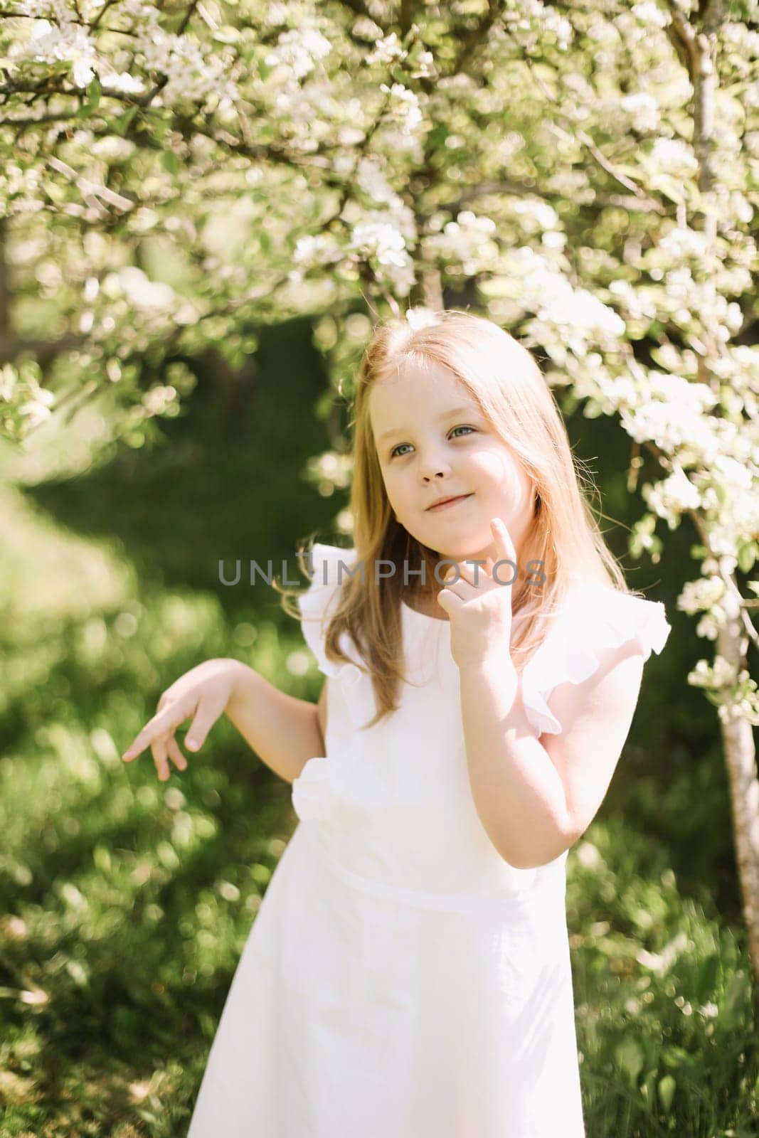 Portrait of little toddler girl in white romantic dress in blossoming garden in spring. Cute beautiful lovely child having fun in a park on a sunny day. by paralisart