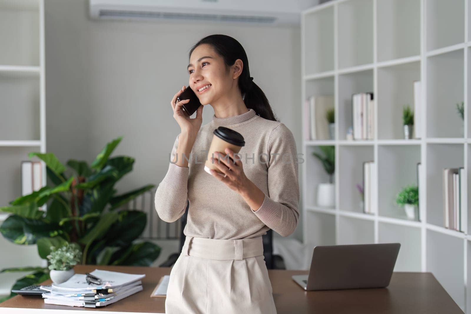 A woman is talking on her cell phone while holding a coffee cup. She is smiling and she is in a good mood. The scene takes place in a room with a desk and a potted plant