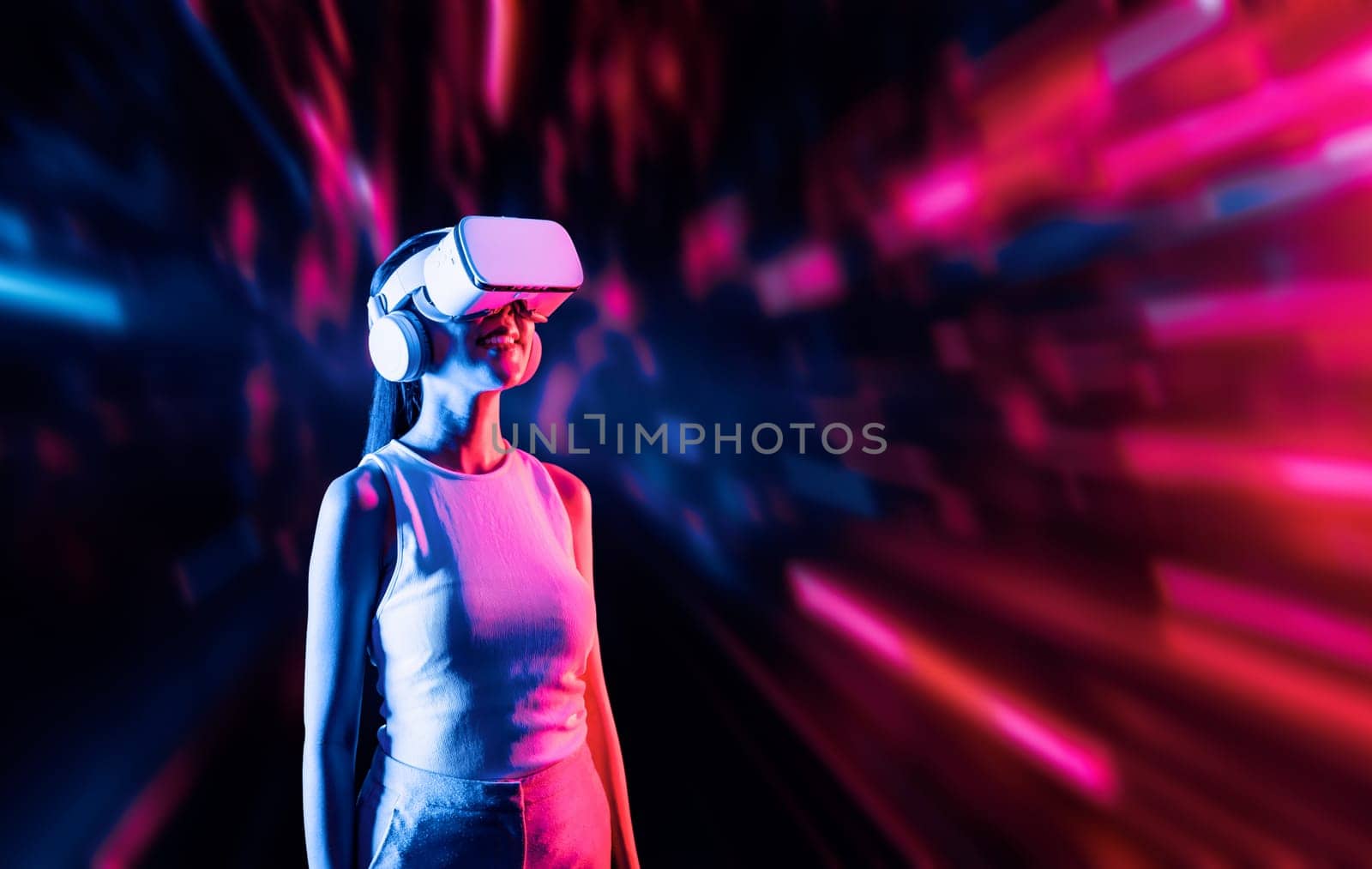 Smart female standing surrounded by neon light wearing VR headset connecting metaverse, future cyberspace community technology. Elegant woman looking faraway and smiling satisfactorily. Hallucination.