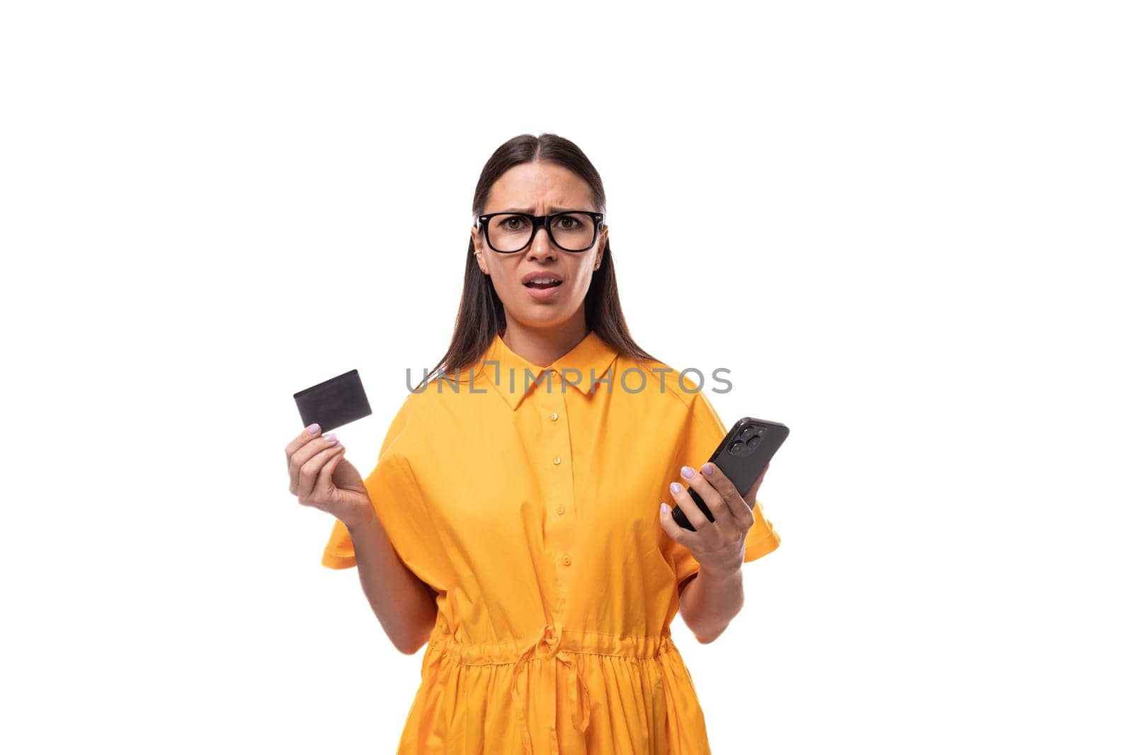 young black-haired woman with glasses dressed in an orange summer dress holds a plastic card and a smartphone and doubts.