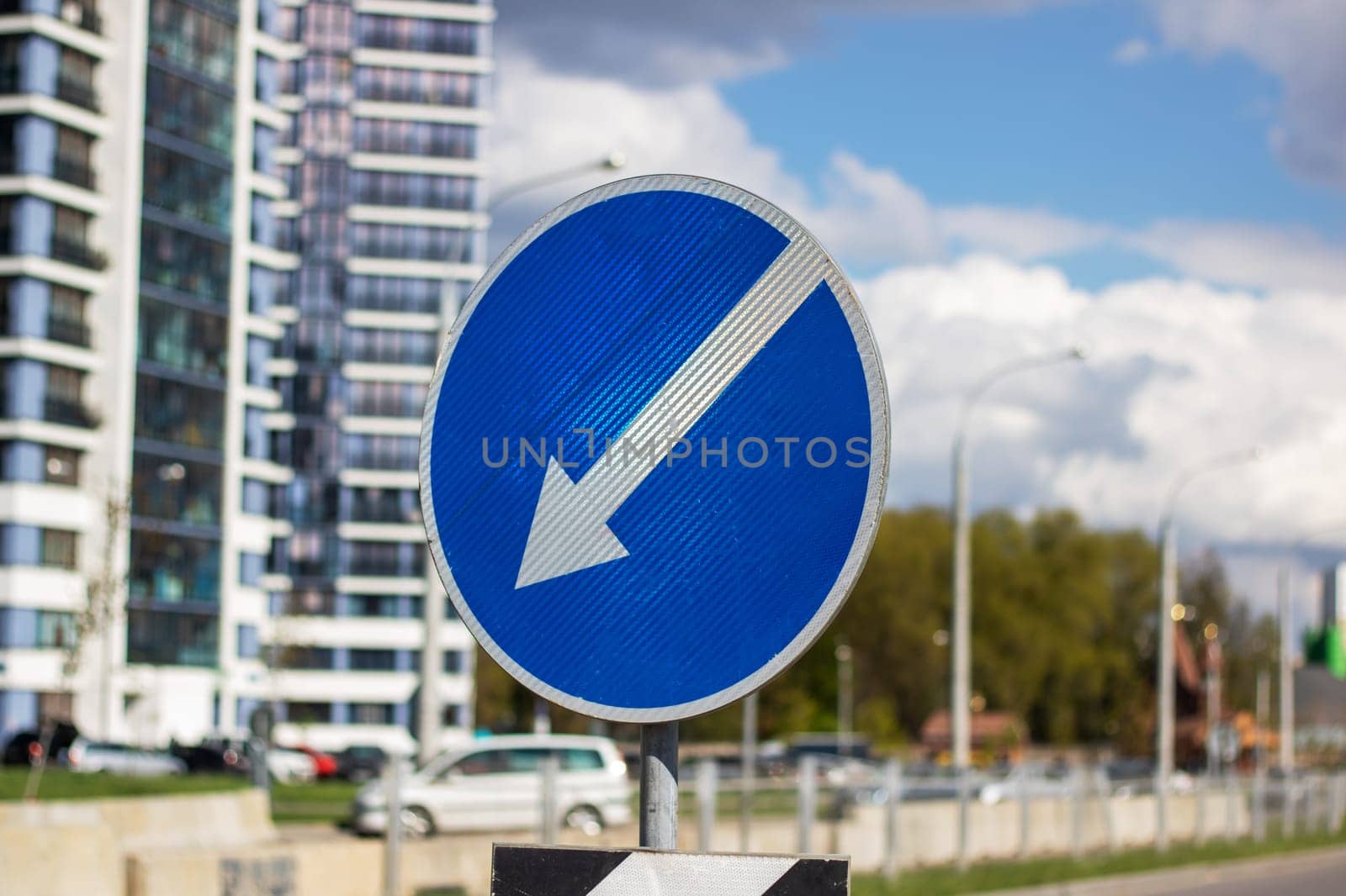 A blue traffic sign with an arrow pointing left against a backdrop of a cloudy sky in the daytime, with skyscrapers, buildings, trees, and plants in the city