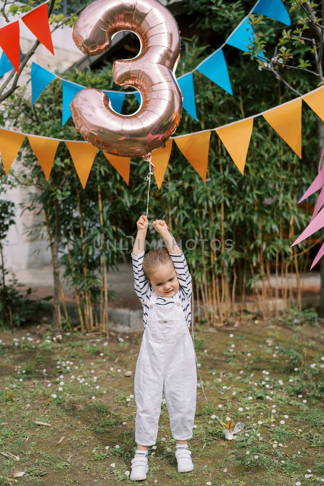 Little girl stands on a green lawn under colorful flags on a string and holds an inflatable number 3 . High quality photo