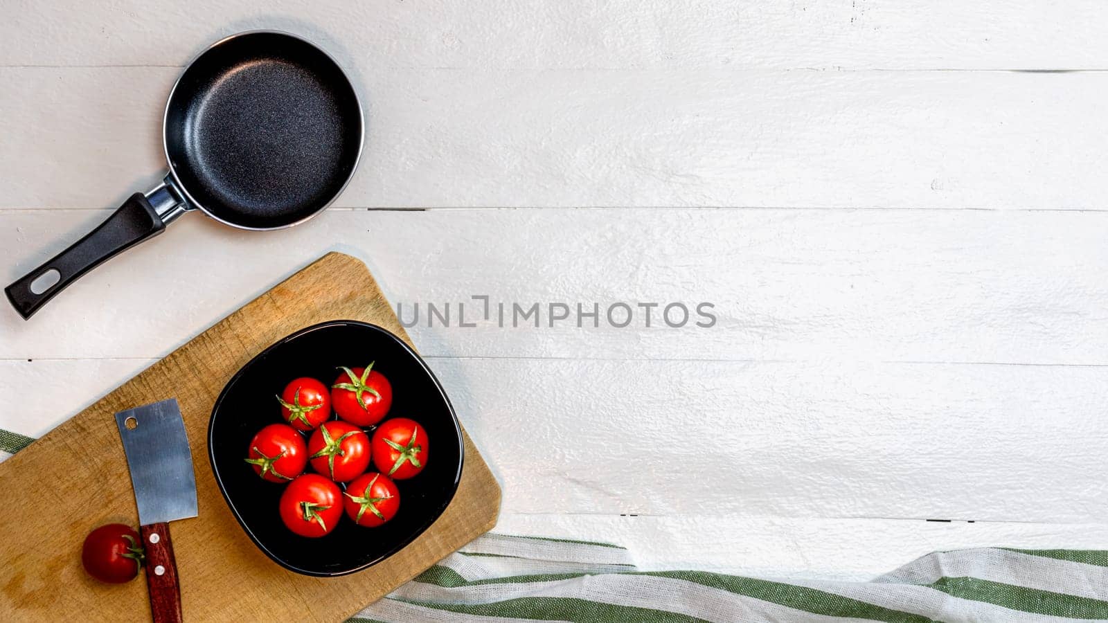 Top view of knife, small pan and fresh ripe cherry tomatoes in small black bowl on a rustic white wooden table. Ingredients and food concept