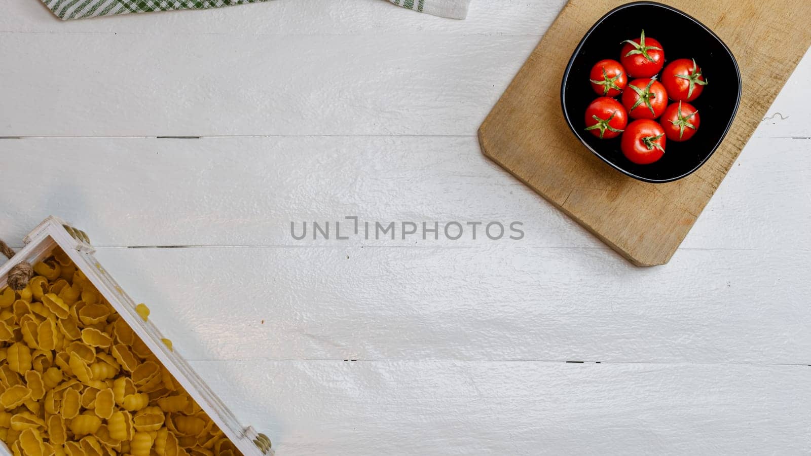 Top view of pasta in a wooden crate and fresh ripe cherry tomatoes in a black bowl on a rustic white wooden table. Ingredients and food concept