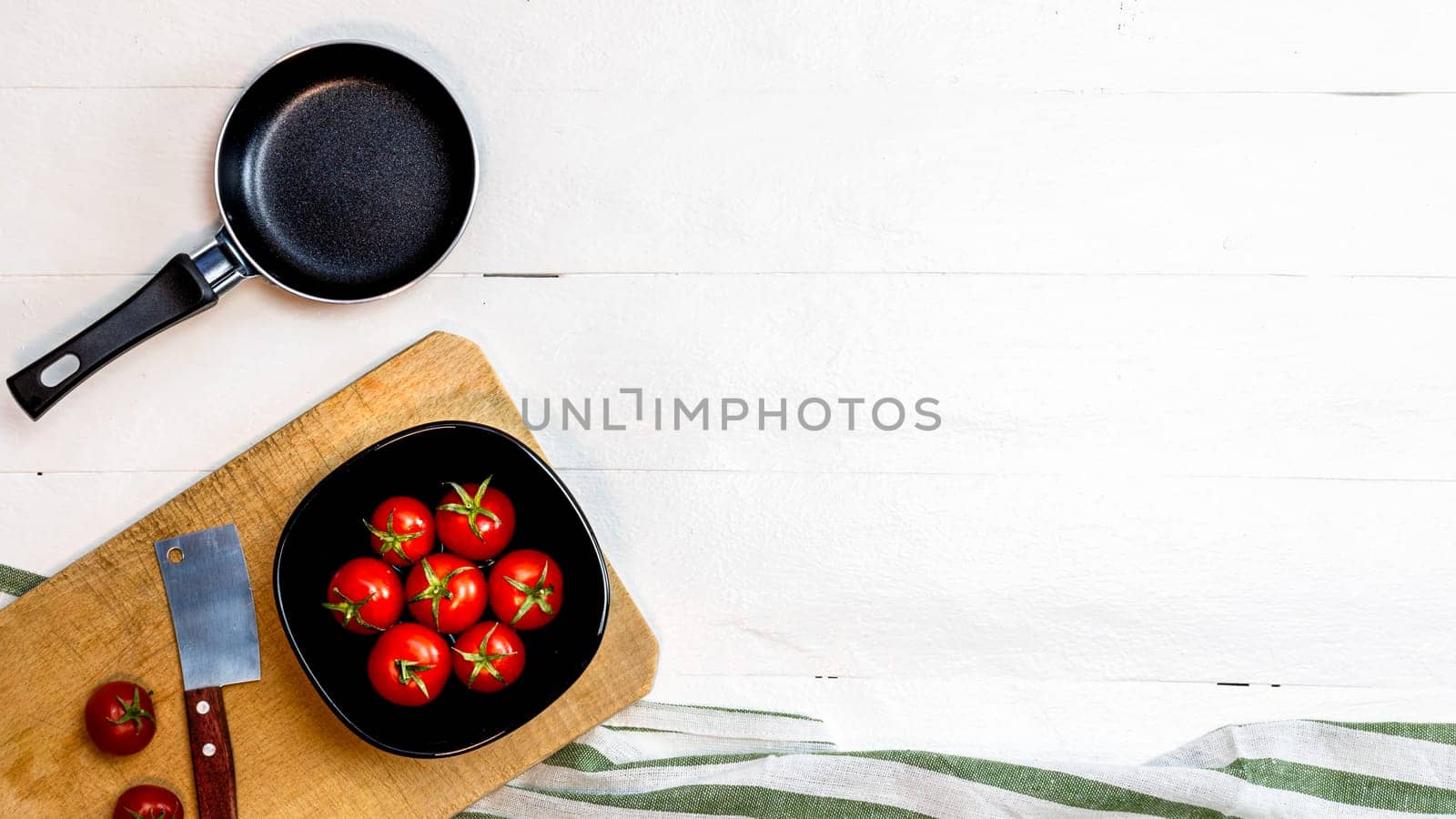 Top view of knife, small pan and fresh ripe cherry tomatoes in small black bowl on a rustic white wooden table. Ingredients and food concept